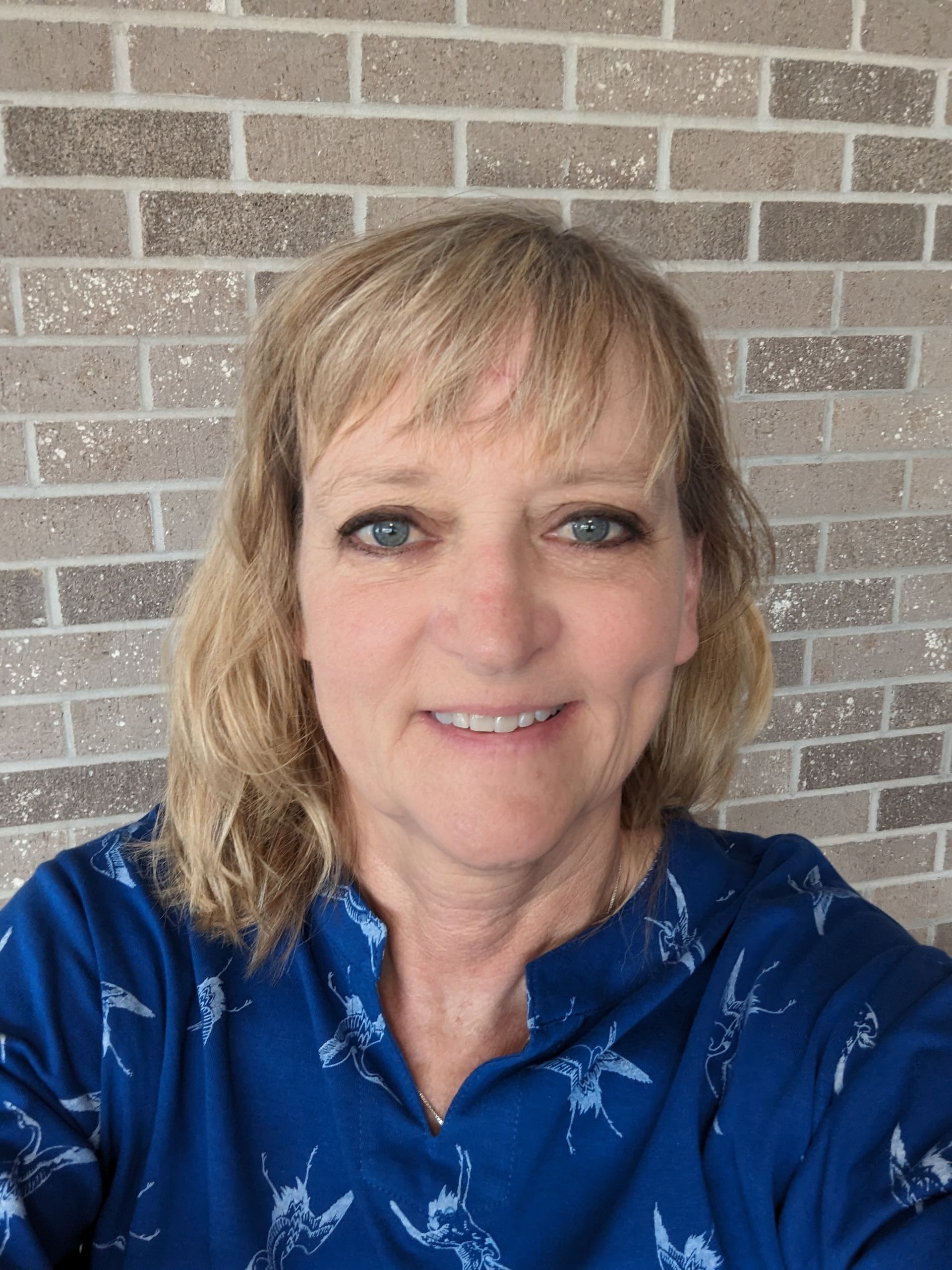 Woman with blonde hair, smiling in front of a brick wall. She wears a blue patterned top.
