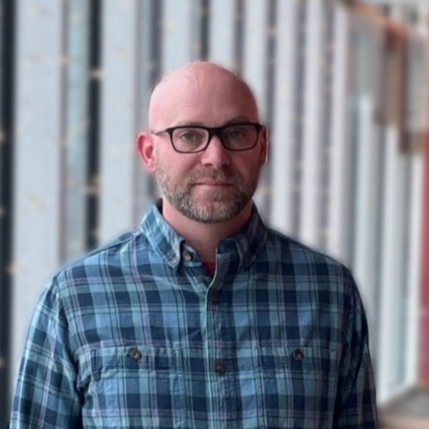 Bald man with glasses wearing a blue plaid shirt, looking at the camera. Neutral background.