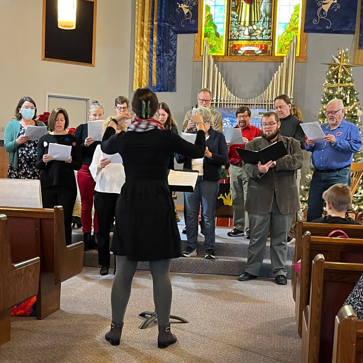 Choir in a church performing, conductor at front. Christmas tree in the background.