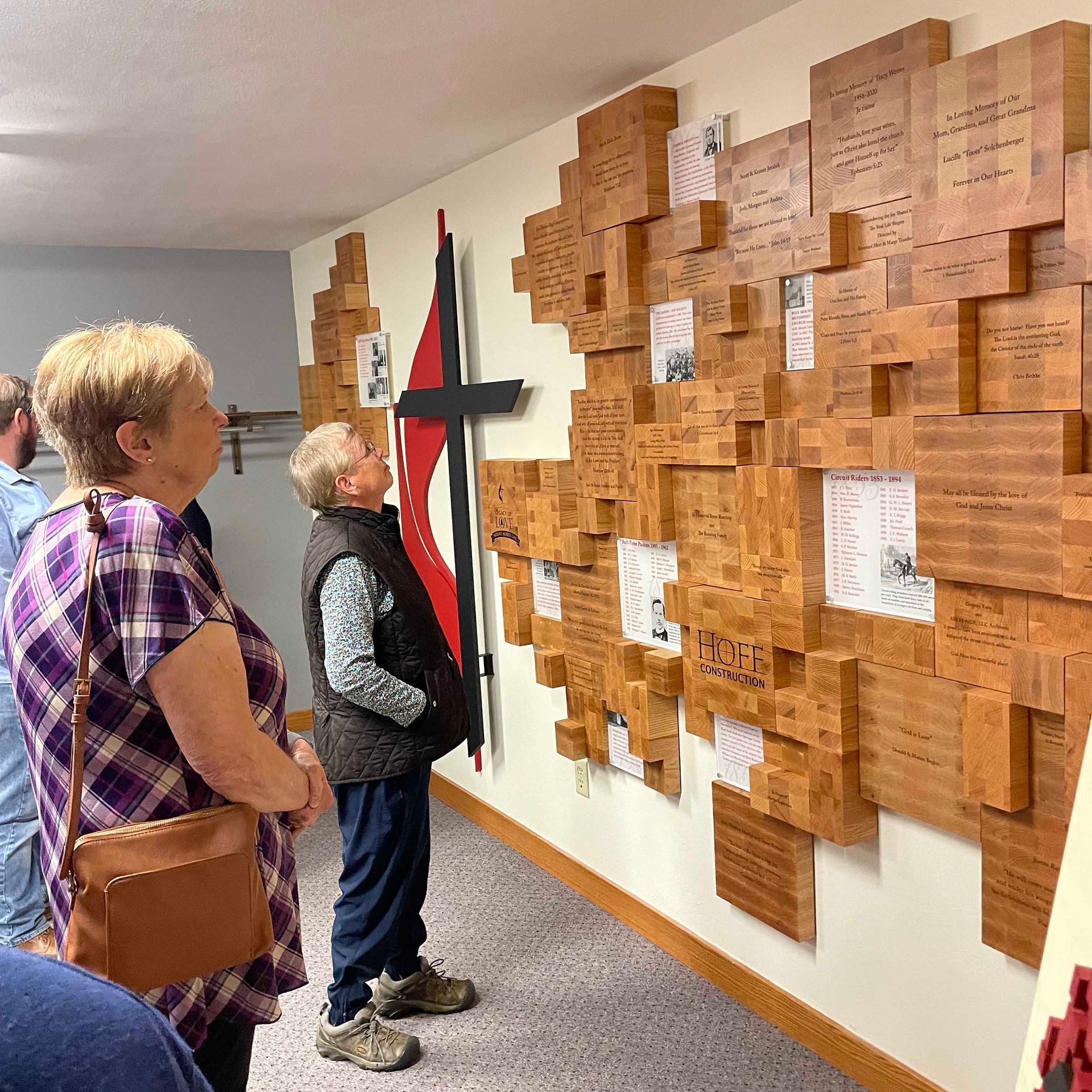 Two people looking at a wall covered with wooden blocks and a large cross, indoors.