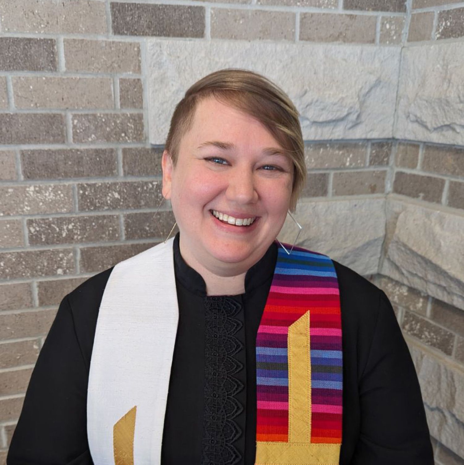 Person in black clerical garb smiles, wearing two stoles, one white, one colorful, standing against a brick wall.