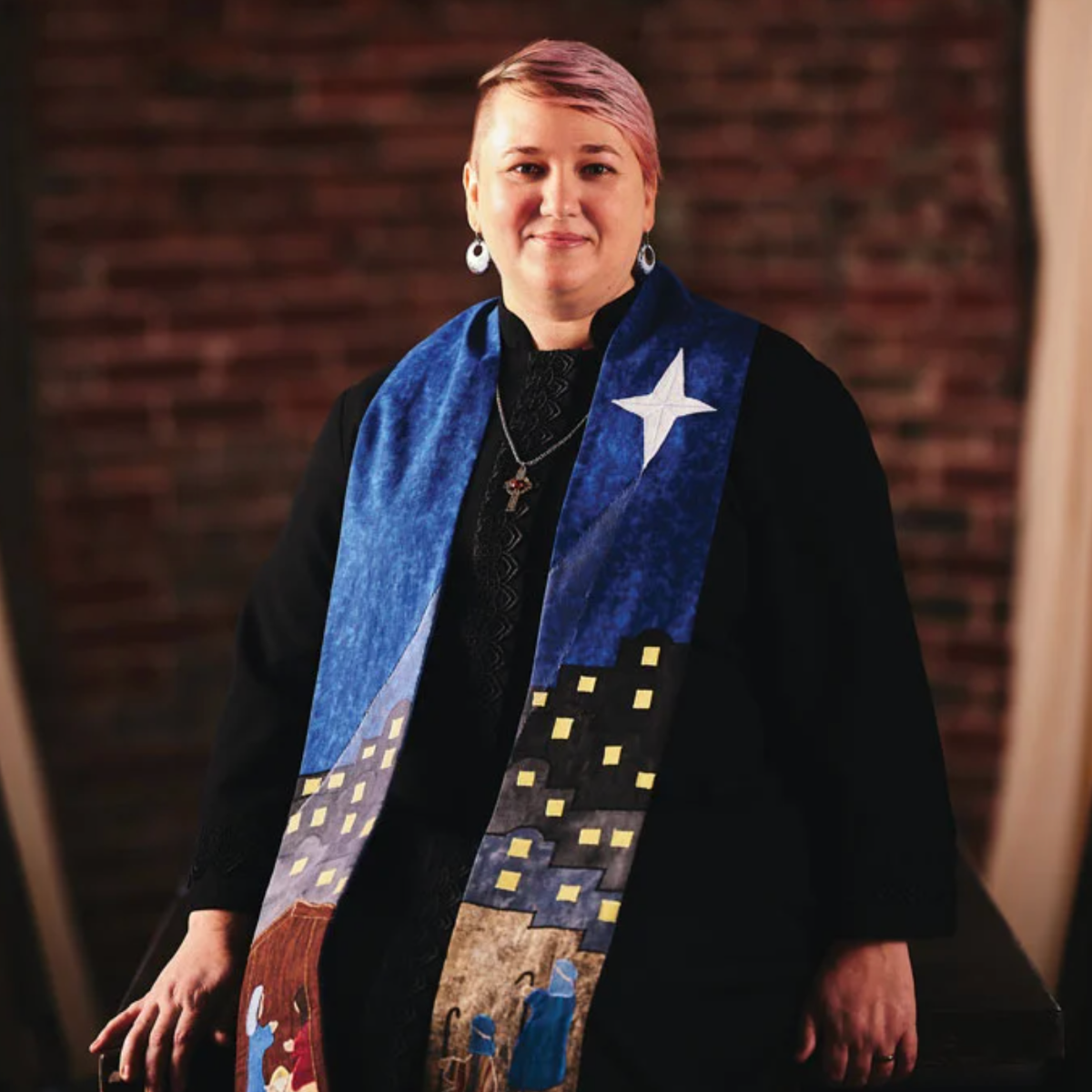 Person in a black robe with a blue, star-adorned stole. They have pink hair, are smiling, and are in a building.