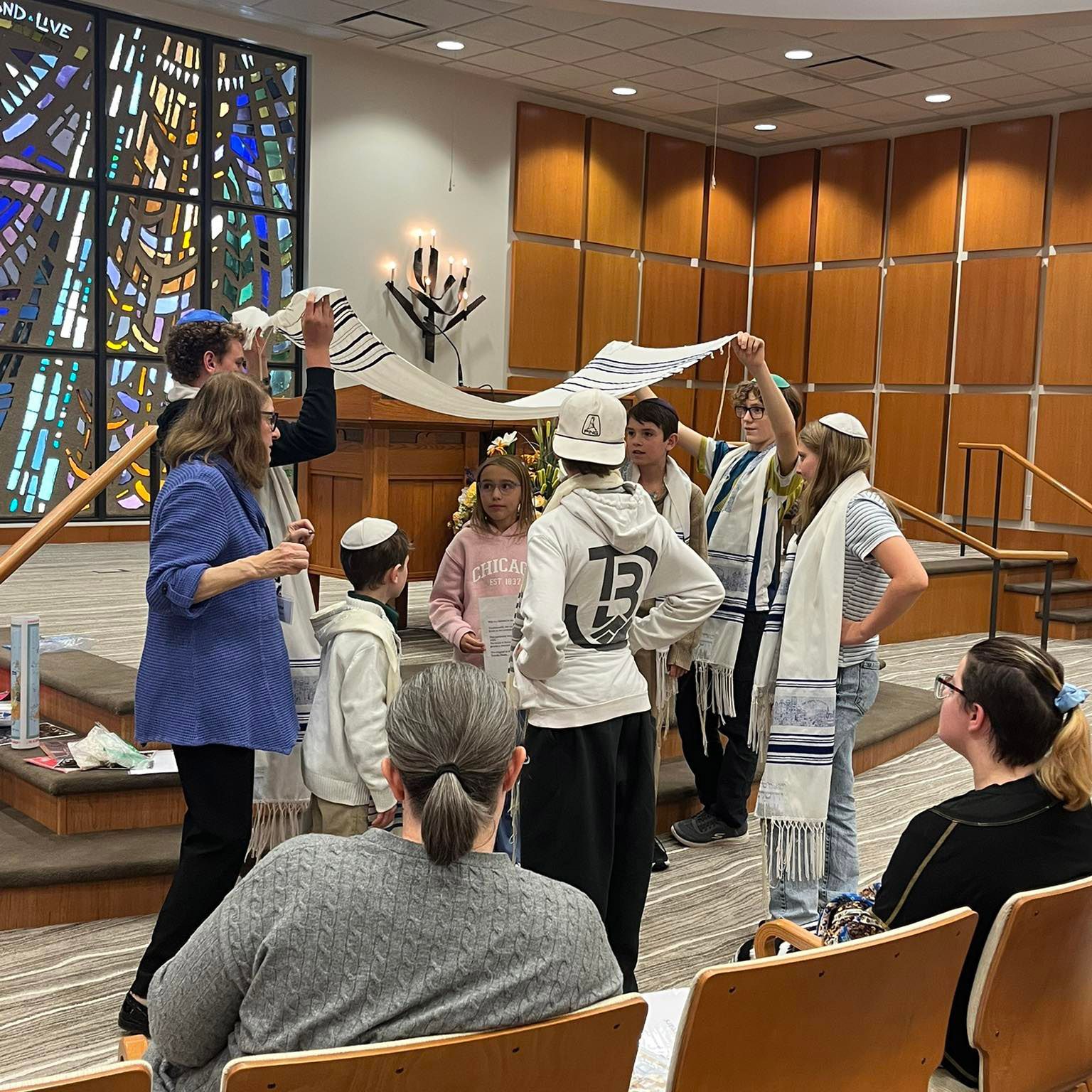 People gathered indoors around a child in a white costume, holding props in a wood-paneled room with stained glass.