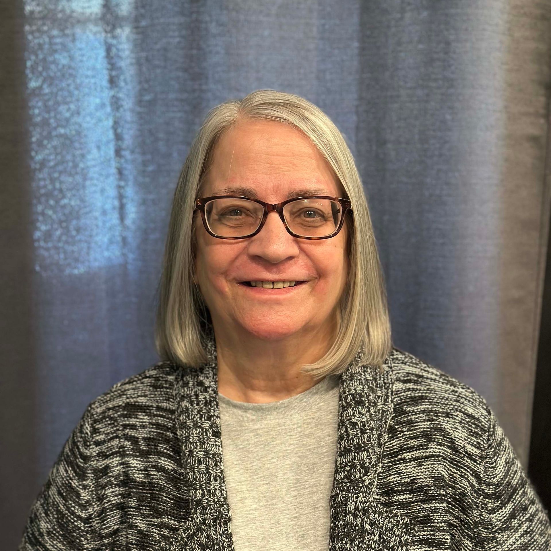 Woman with glasses smiling, gray hair, wearing gray sweater and patterned cardigan, blue curtain backdrop.