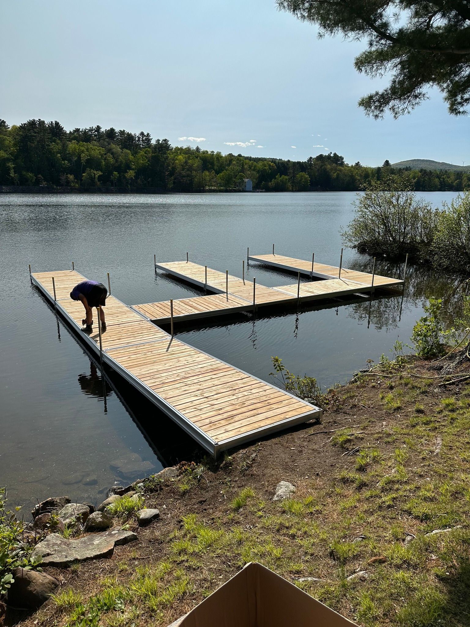 Floating docks on a calm lake beside a grassy shore, with trees and a blue sky in the background