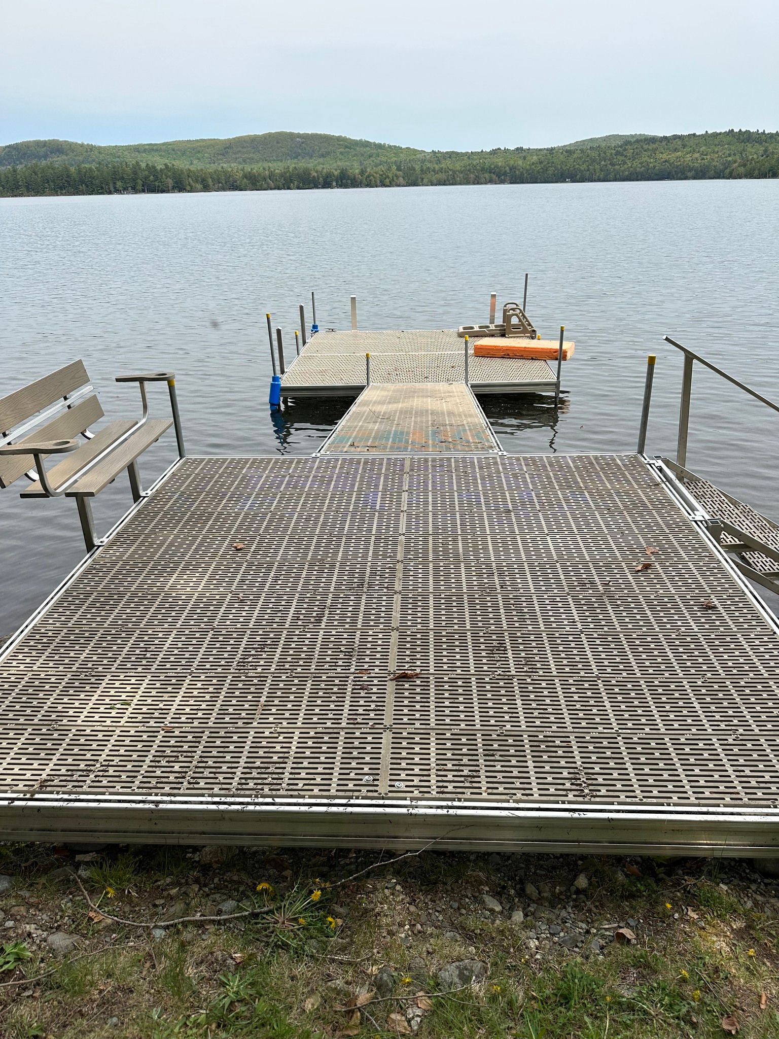 Damaged dock with bent metal grating extending into a calm lake, with a wooded shoreline in the distance