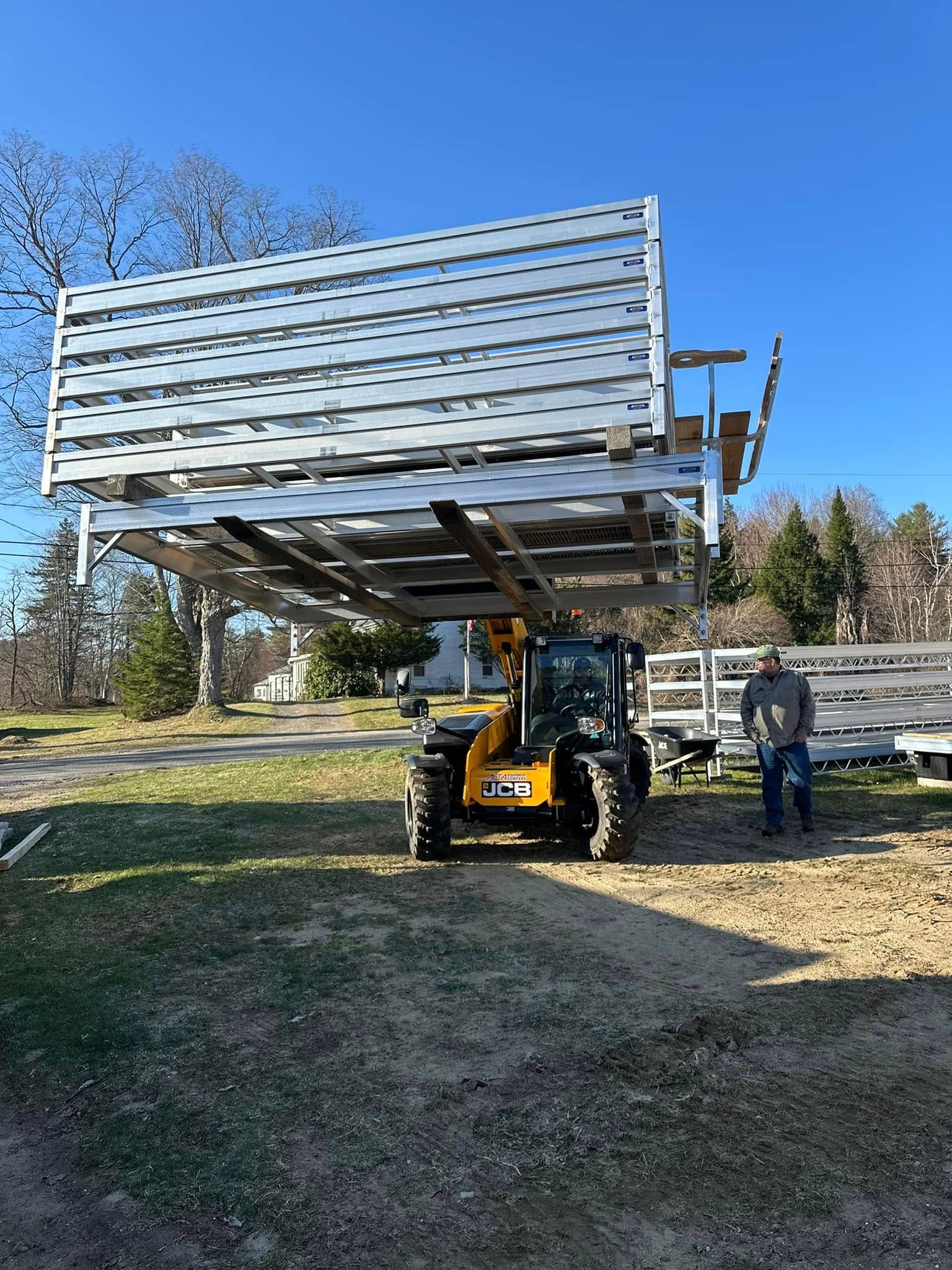 Skid steer lifting a large metal platform with a person standing beside it outdoors.