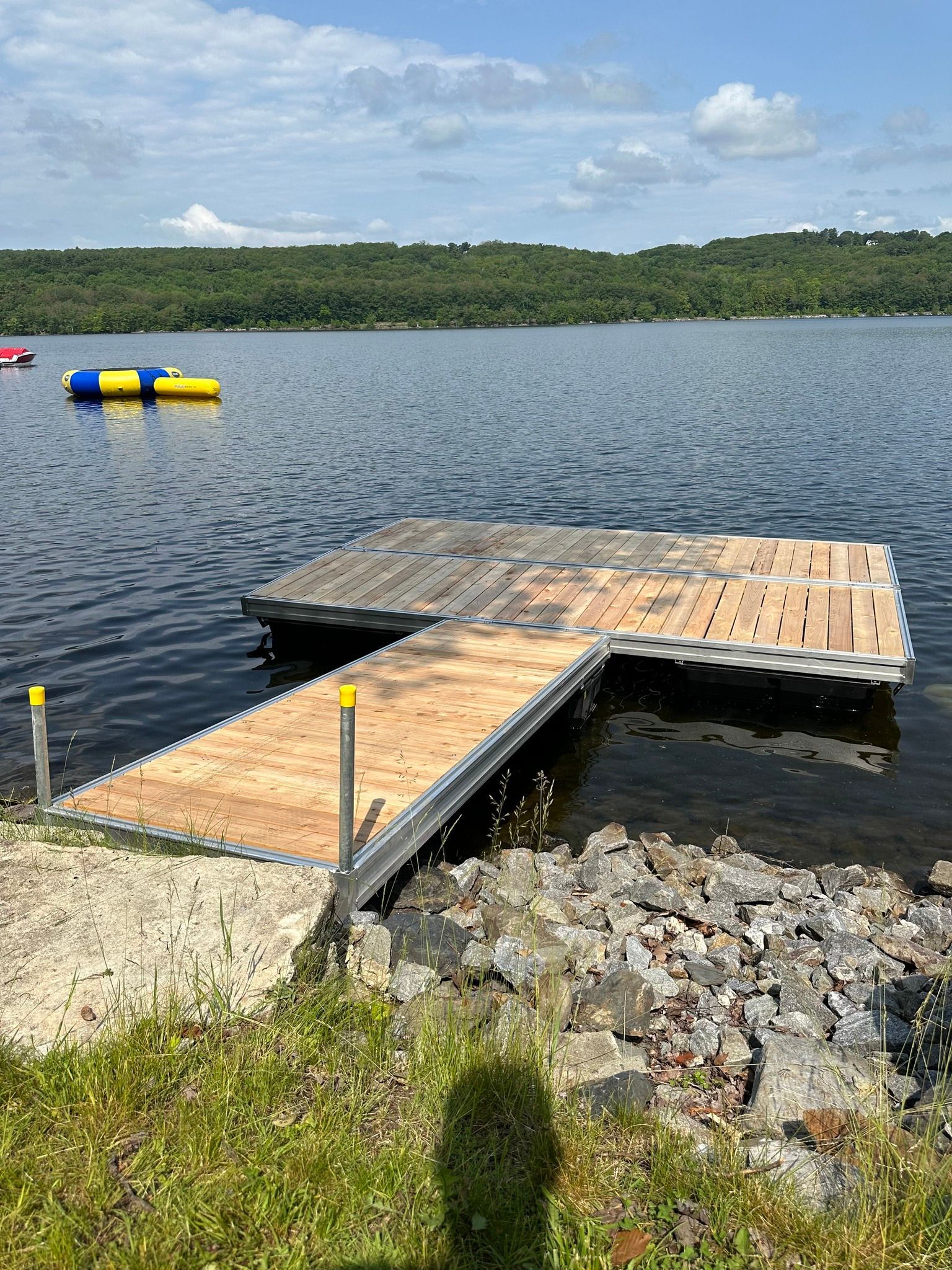 Floating dock on a lake with yellow and blue water floats, rocky shore, and forested shoreline in the distance