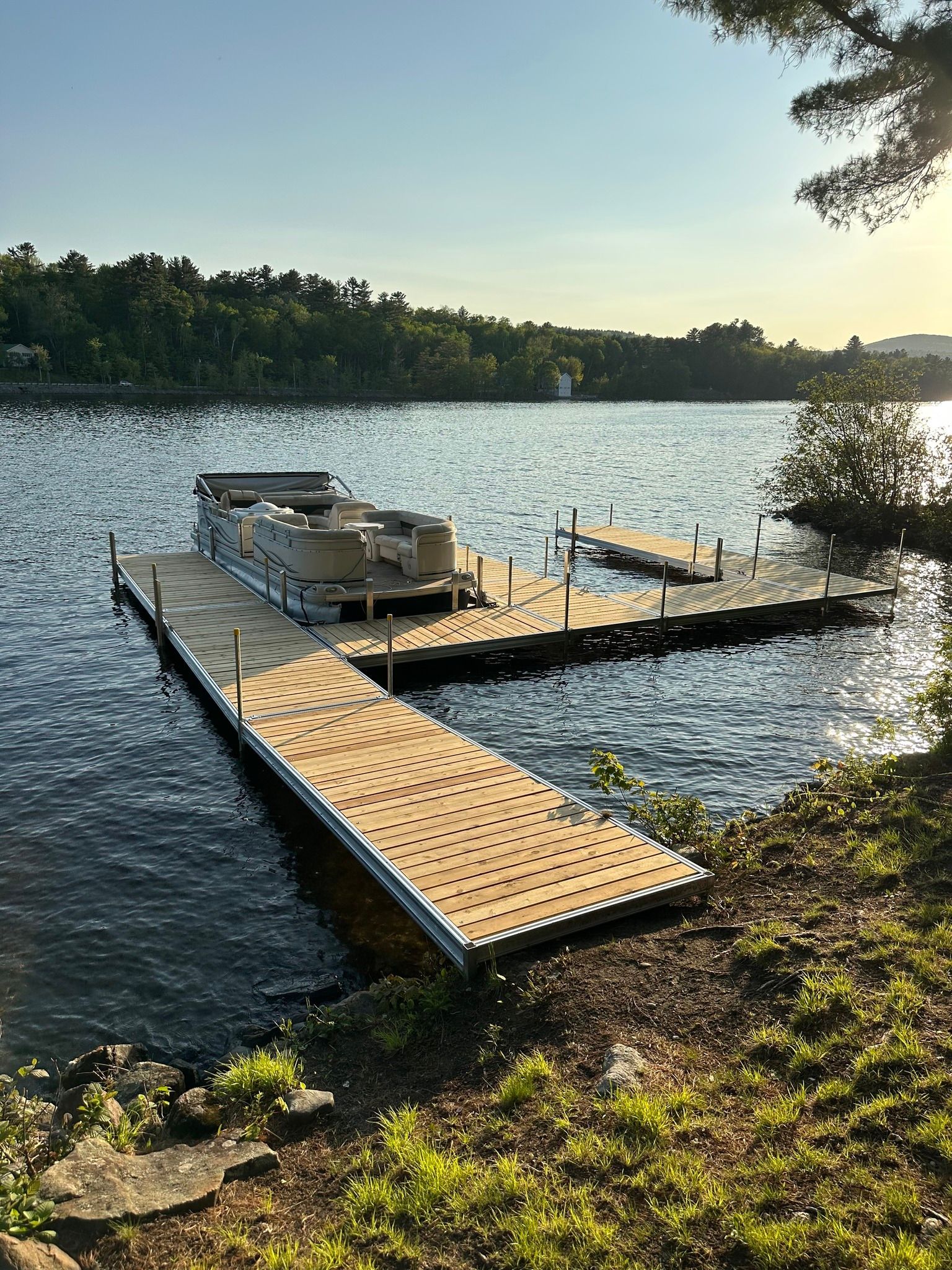 Wooden dock and pontoon boat on a calm lake at sunset, with trees along the shore.