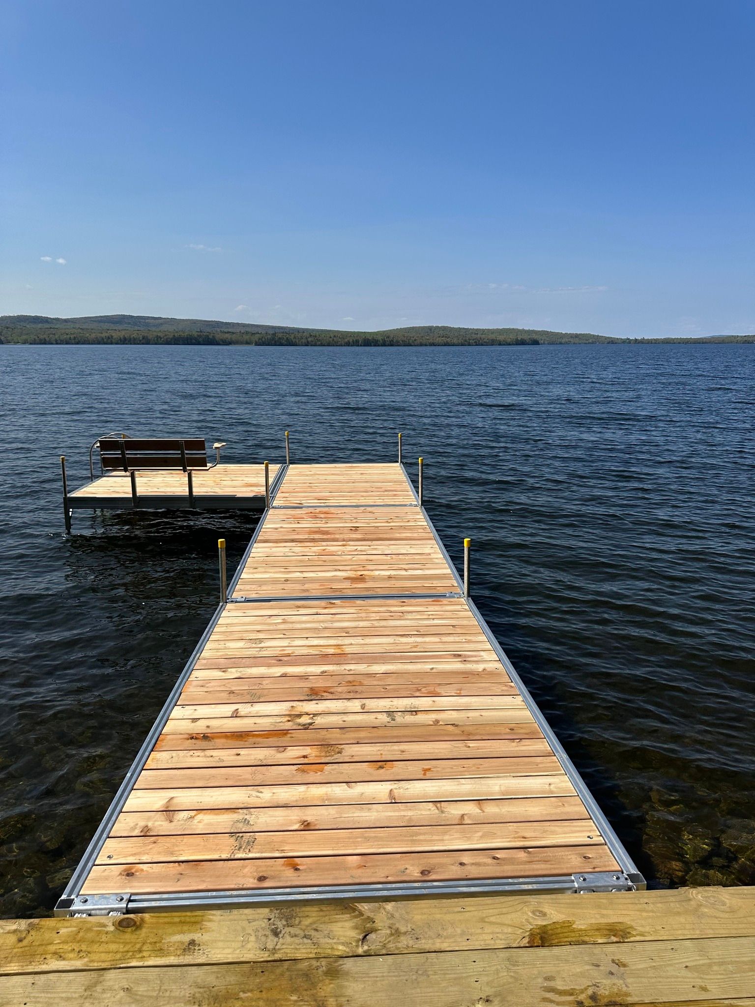 Wooden dock extending into a calm lake under a clear blue sky, with a small bench on the left side.