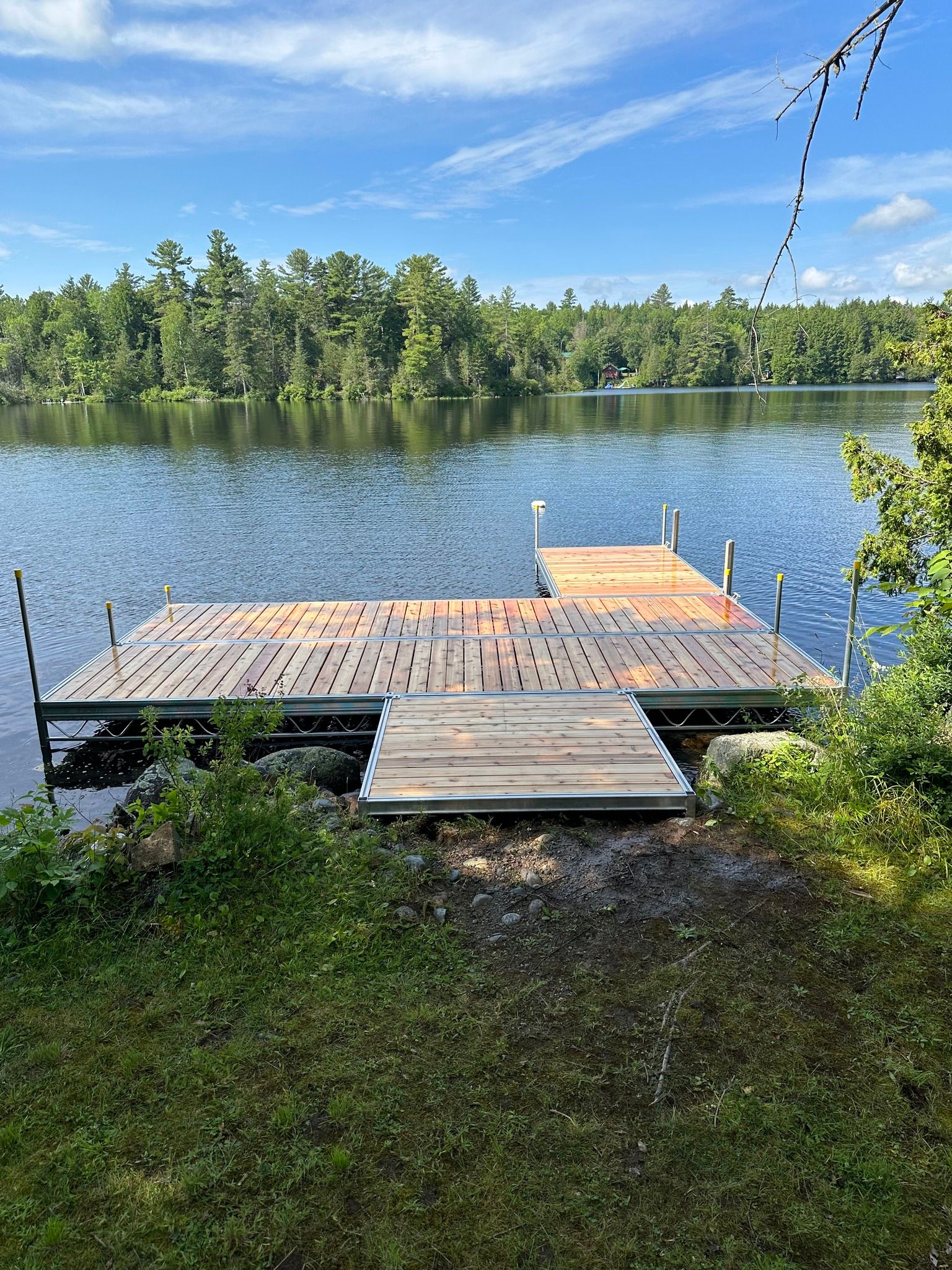 Wooden dock with a floating swim platform on a calm lake, surrounded by green trees and blue sky.