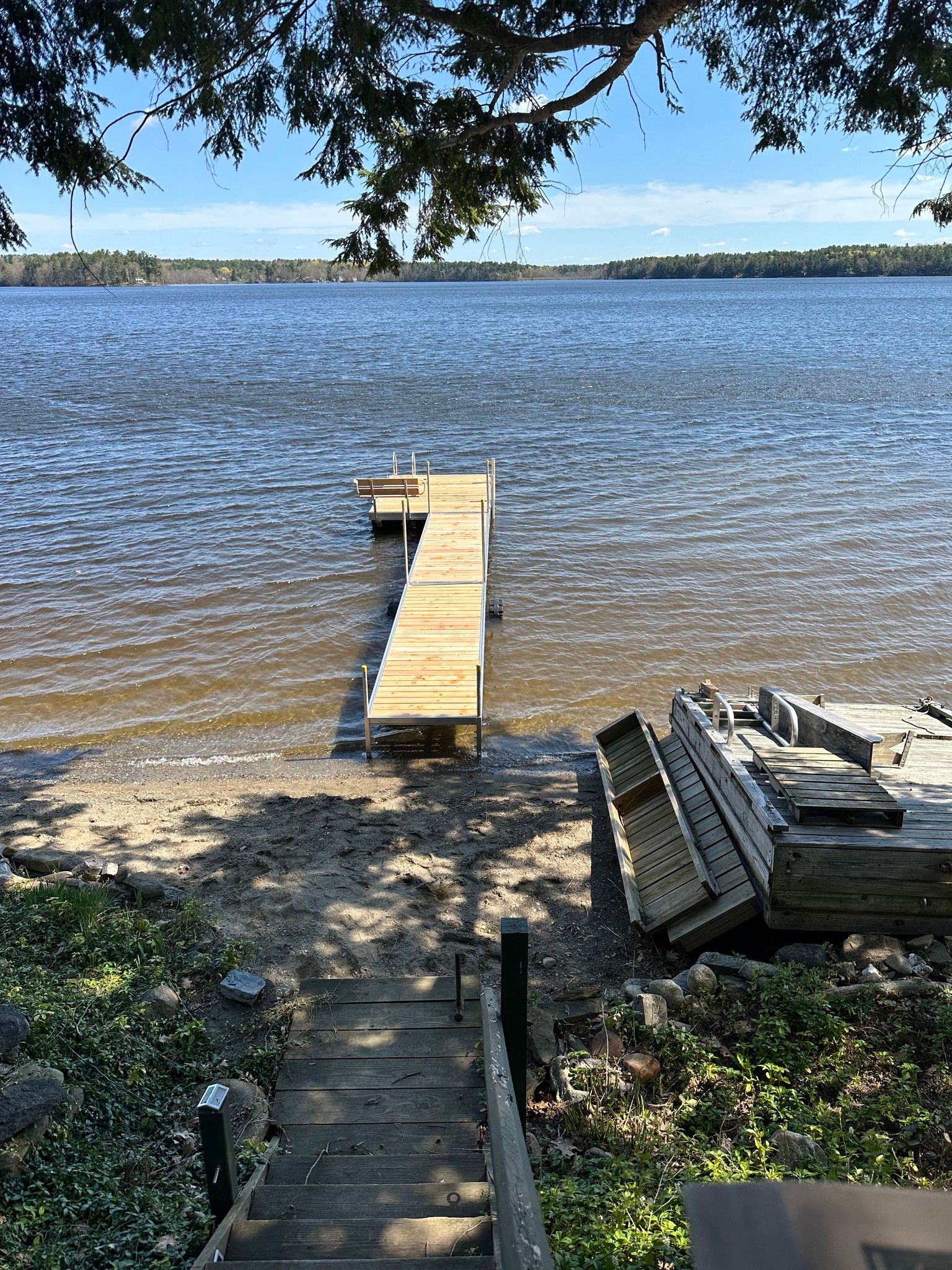 Wooden dock extending into a calm blue lake, with stacked boards and a shaded shoreline in the foreground.