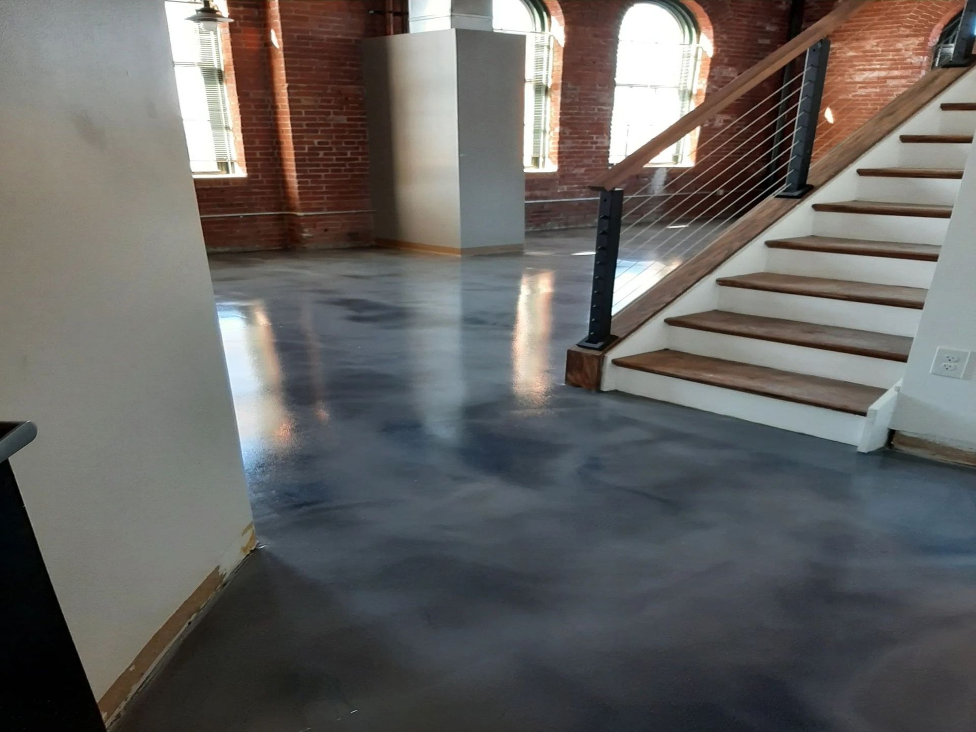 Open-concept interior with concrete floors, stairs with cable railing, brick walls, and arched windows.
