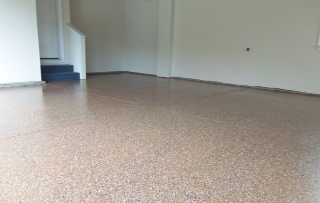 An empty garage with a speckled epoxy floor. Walls are light beige, and a staircase is visible.