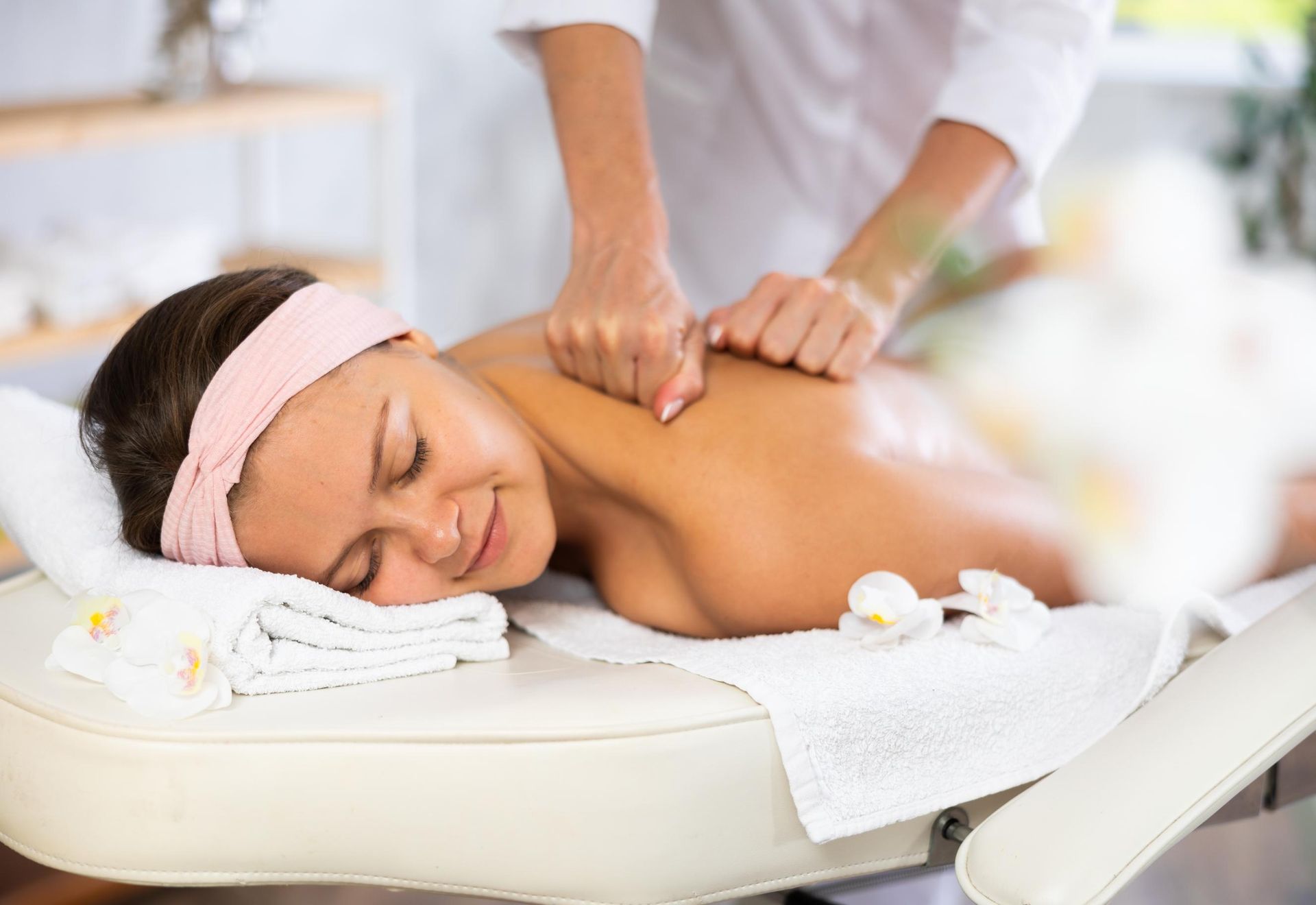 Woman receiving a back massage on a spa table, eyes closed, with white towels and a pink headband.