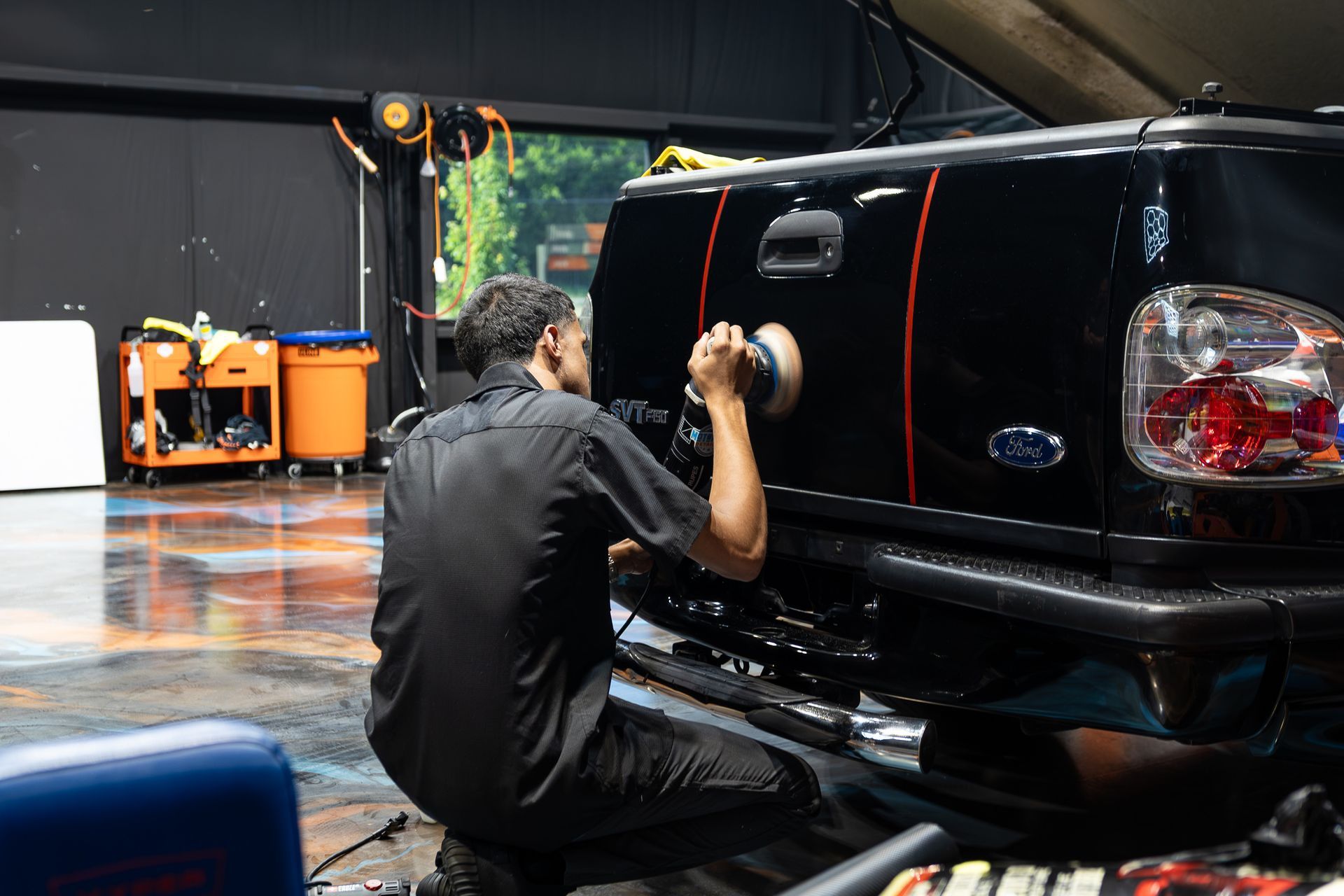 Person polishing a black pickup truck in a garage with orange equipment in the background