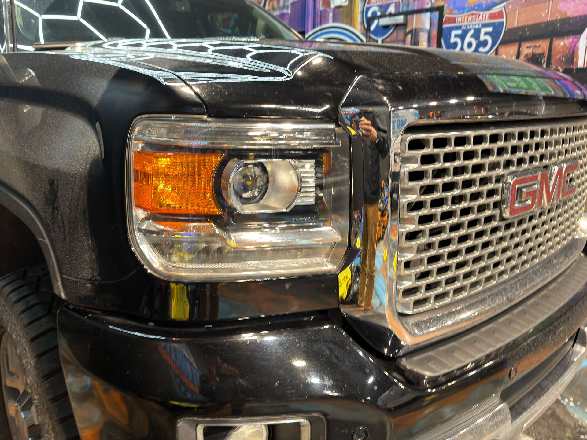 Close-up of a black GMC truck’s chrome grille and headlight at night with colorful mural lights behind it
