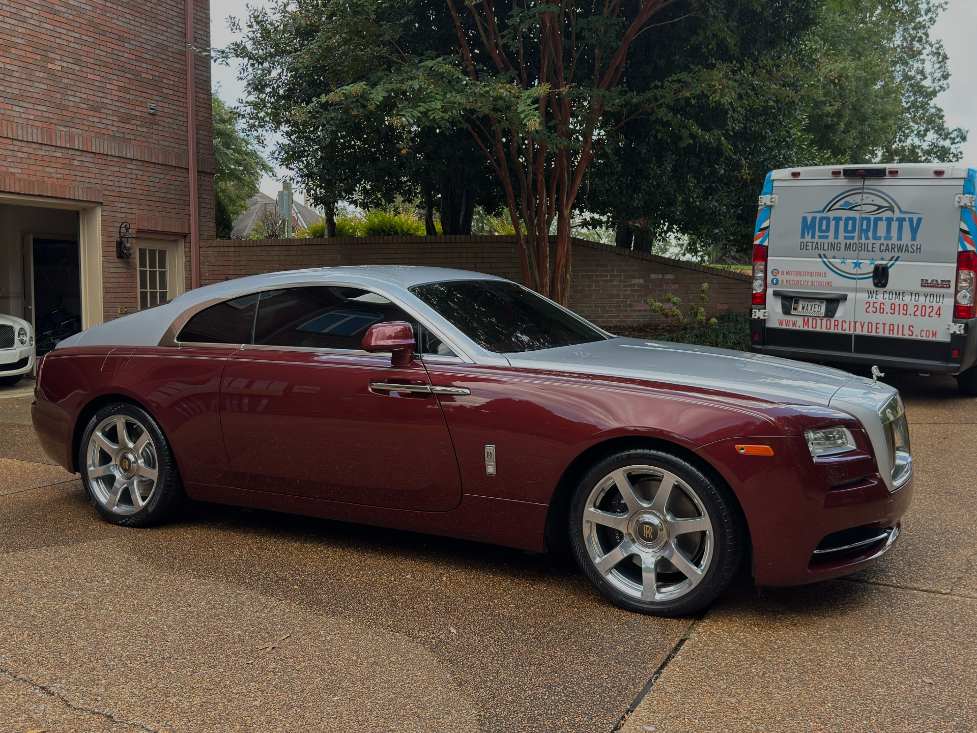 Maroon and silver Rolls-Royce coupe parked in front of a brick building. A van is parked to the right.