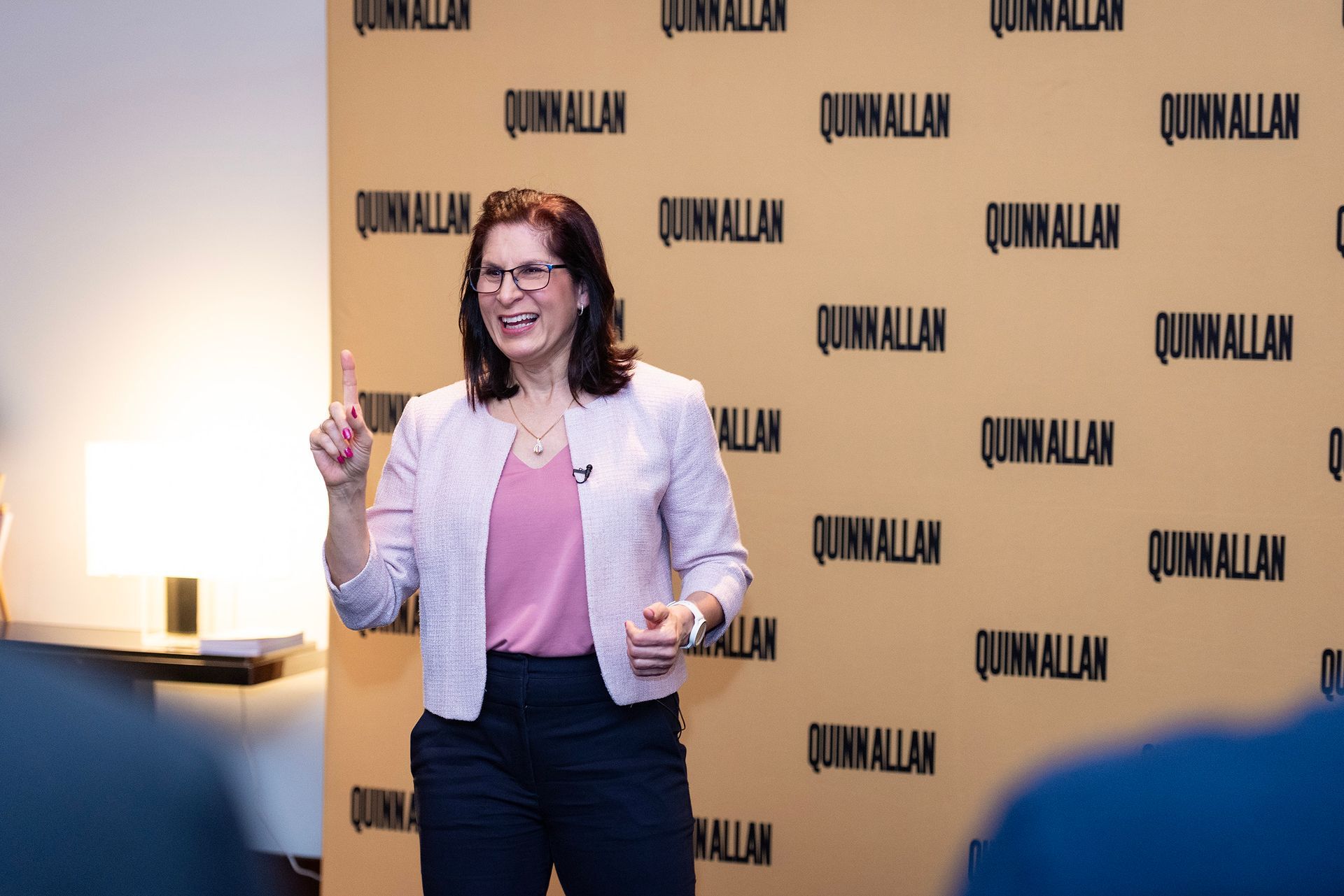 Woman speaking onstage with a microphone, in front of a branded wall with repeated black text.