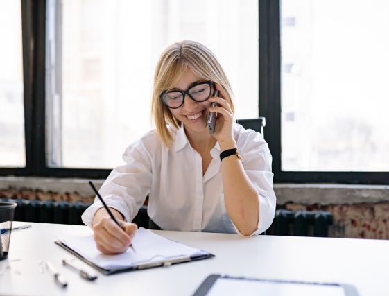Smiling woman in glasses writing on a clipboard and talking on the phone at a bright office desk