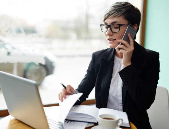 Professional working at a laptop, speaking on a phone and reviewing papers at a desk in a bright office.