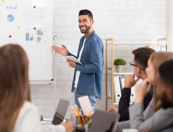 Presenter speaking beside whiteboard in a bright meeting room, with seated attendees listening.