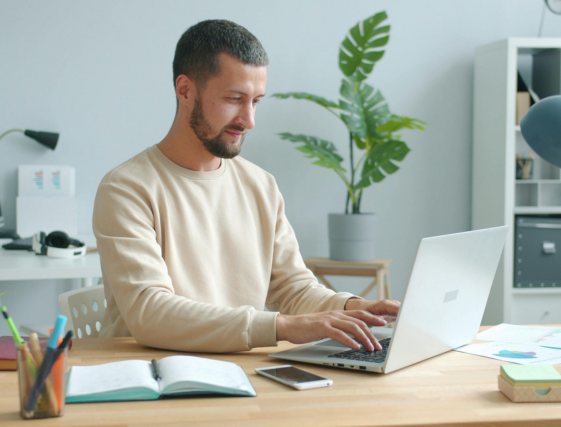 Person working on a laptop at a desk in a bright office, with notebook, phone, and plant nearby