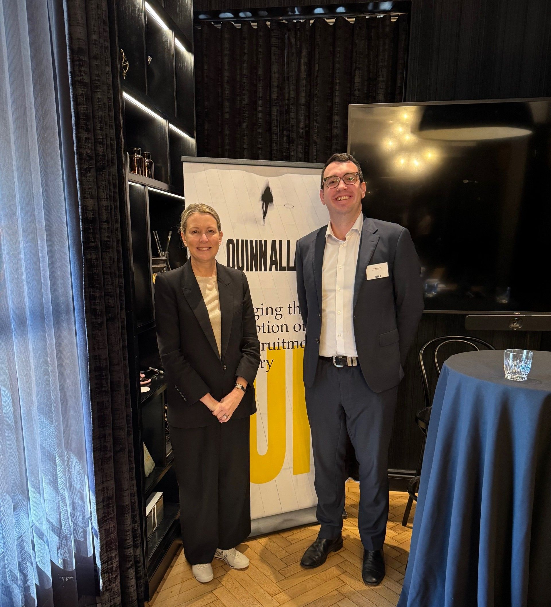 Two people in business attire standing beside a Quinn Allan banner at an indoor event.