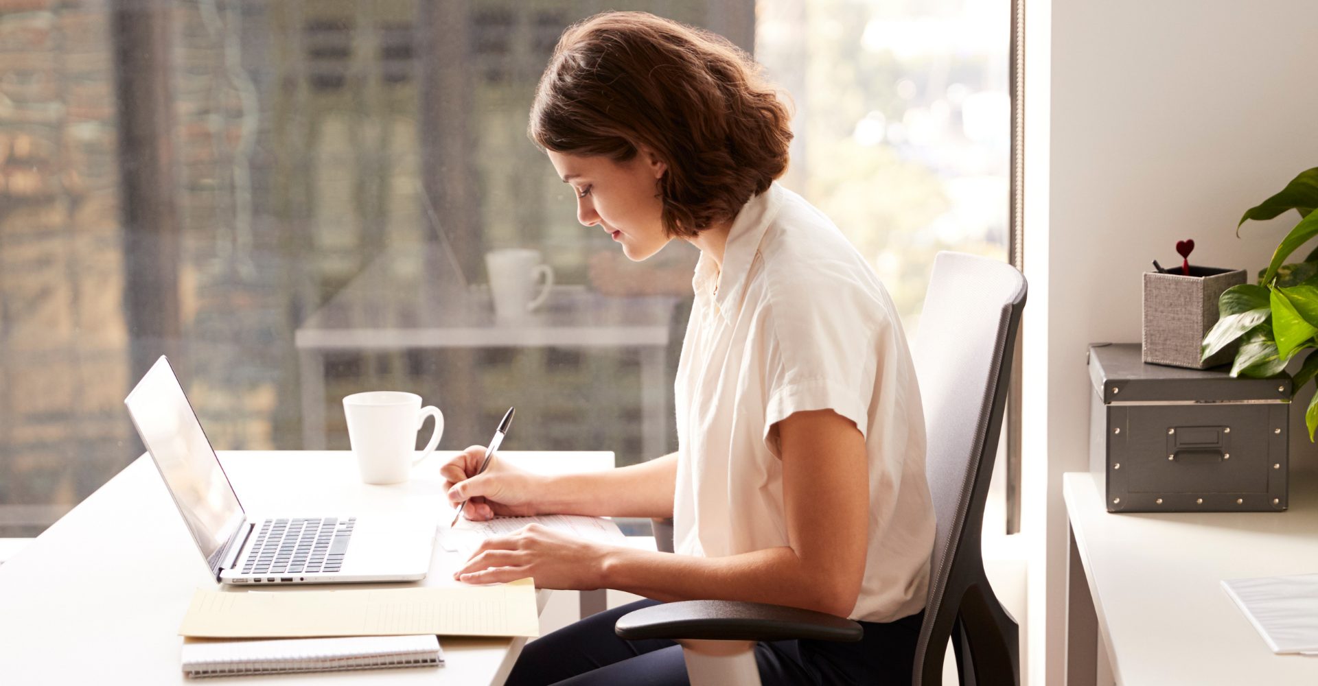 Person working on a laptop at a desk by a sunlit window, with a mug and plant nearby.