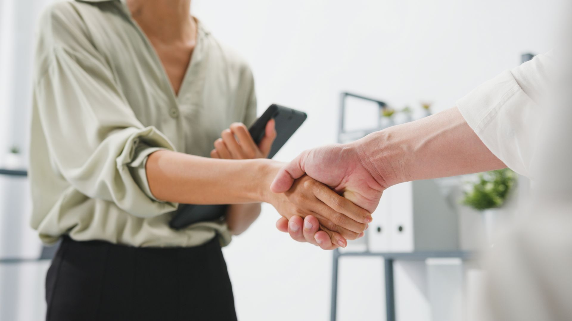 Business handshake in a bright office, with a person holding a smartphone nearby.