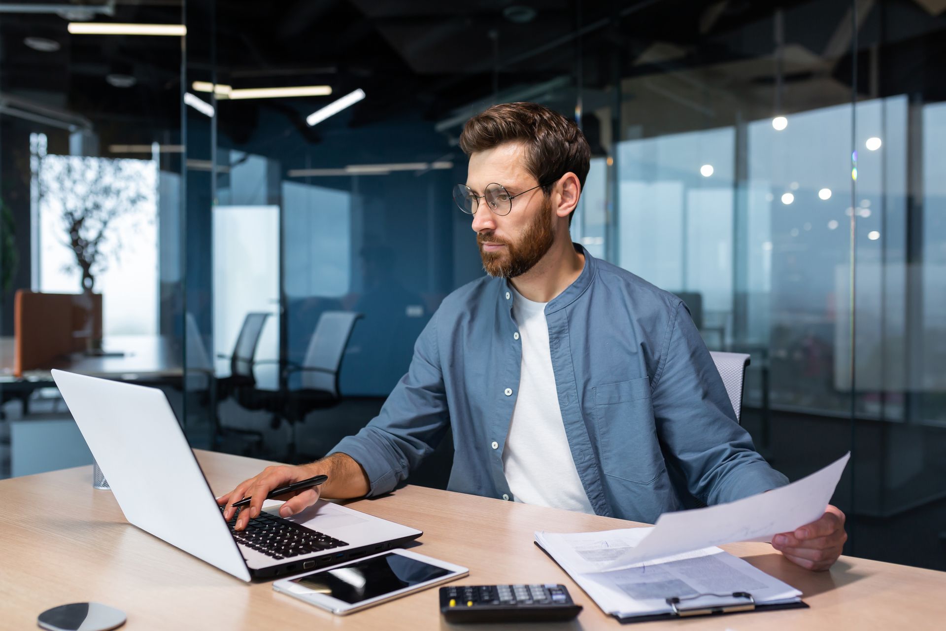 Man working at a desk in a glass office, using a laptop and reviewing papers.