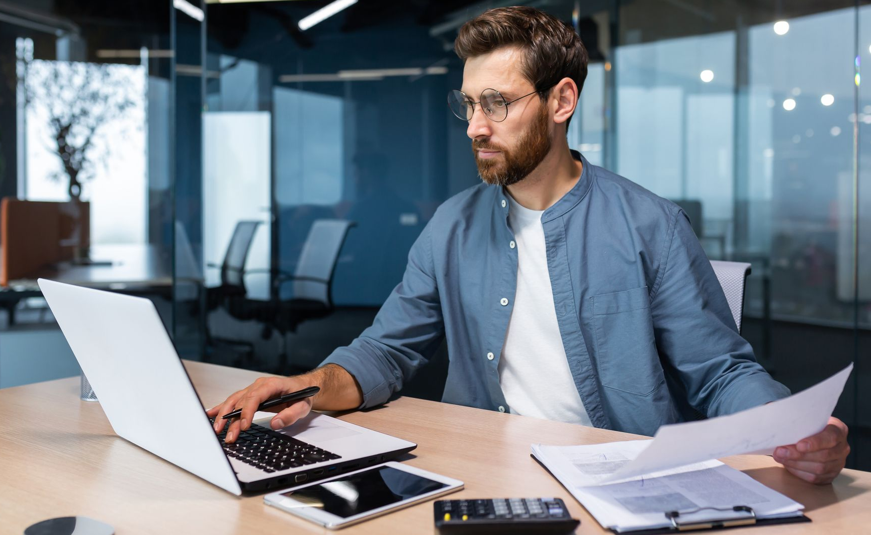 Man working at a desk with a laptop and papers in a modern office, focused on the screen.