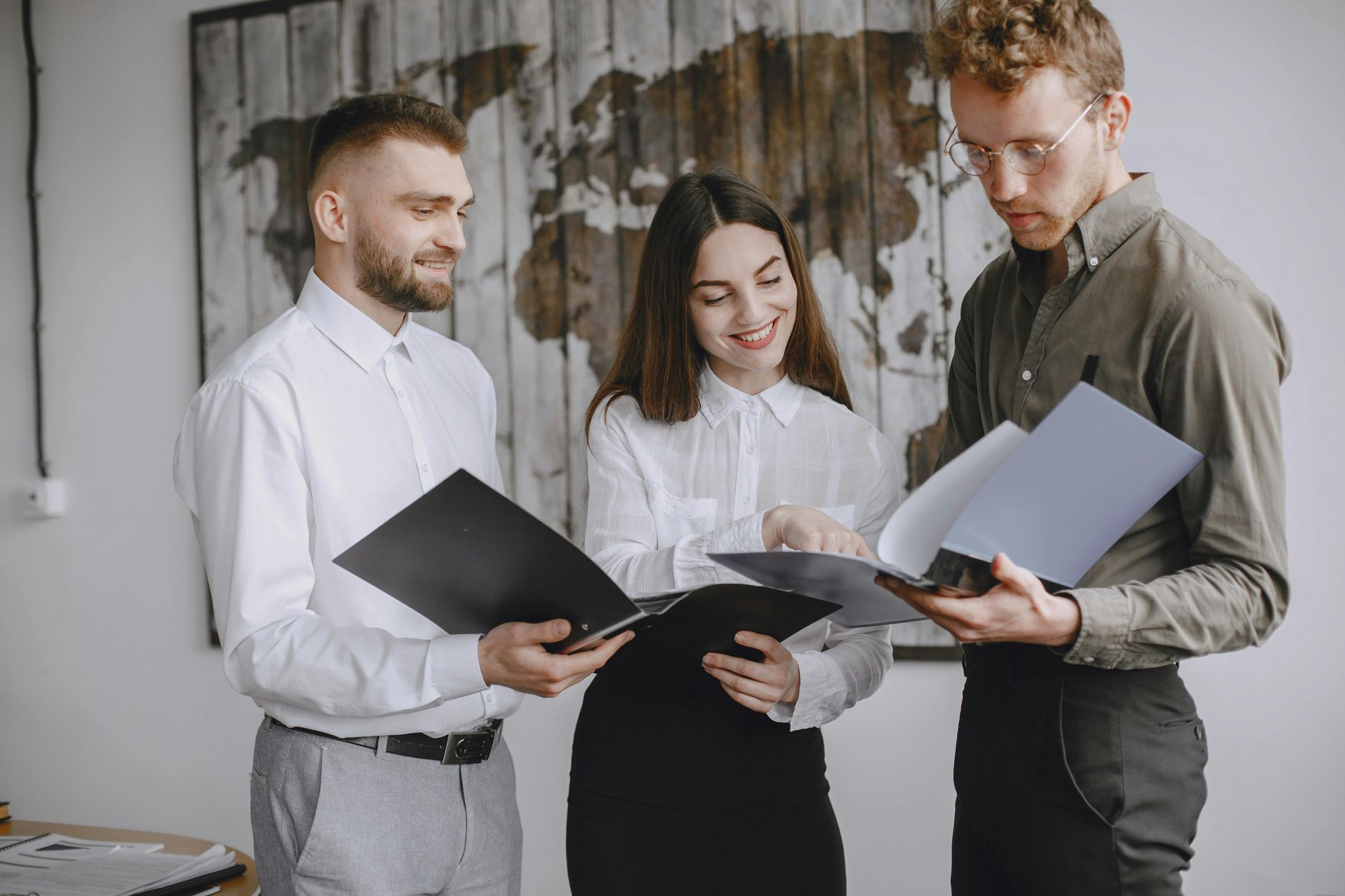 Three coworkers reviewing documents together in a bright office, standing in front of a textured wall.