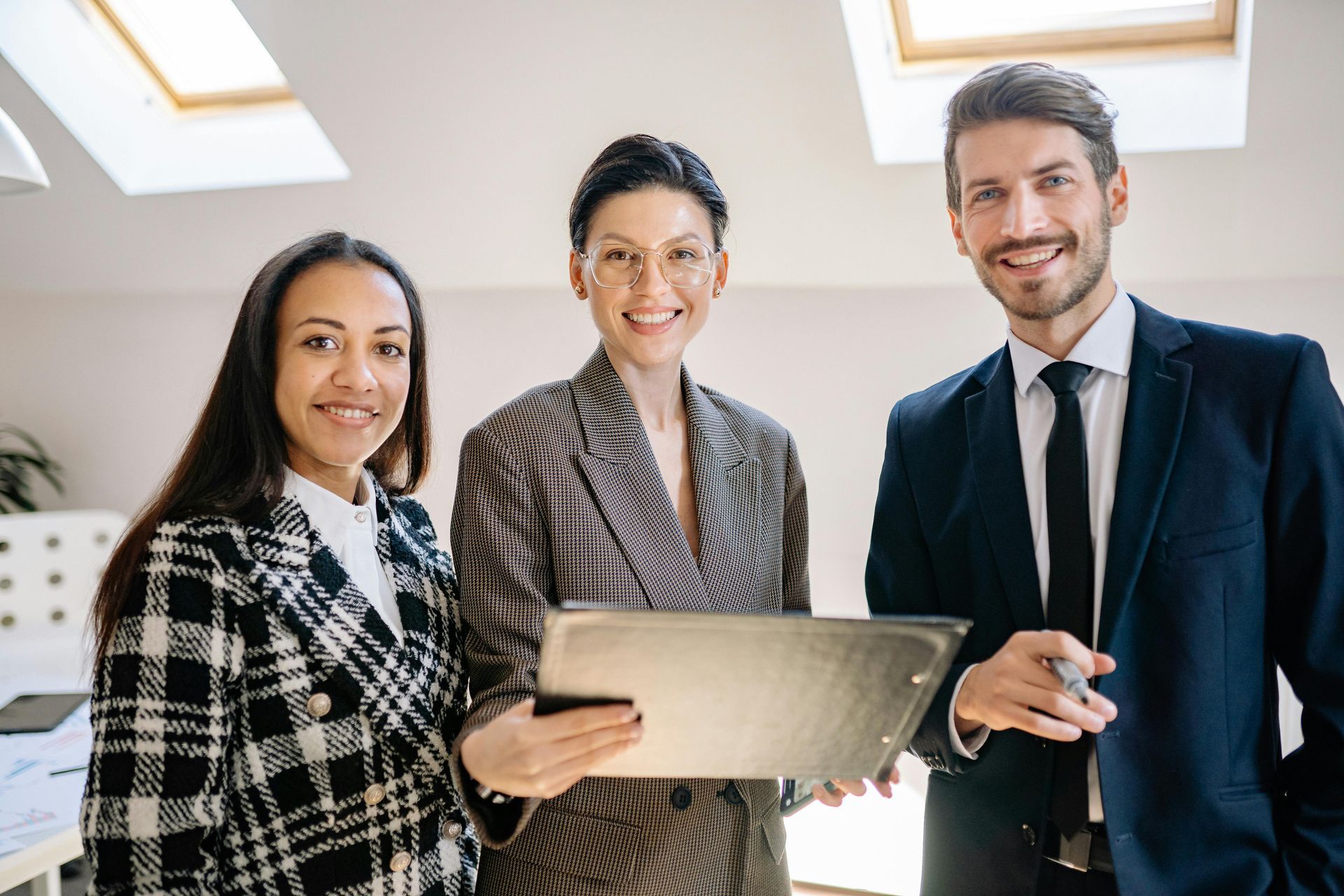 Three professionals in business attire standing indoors, holding a tablet and smiling.