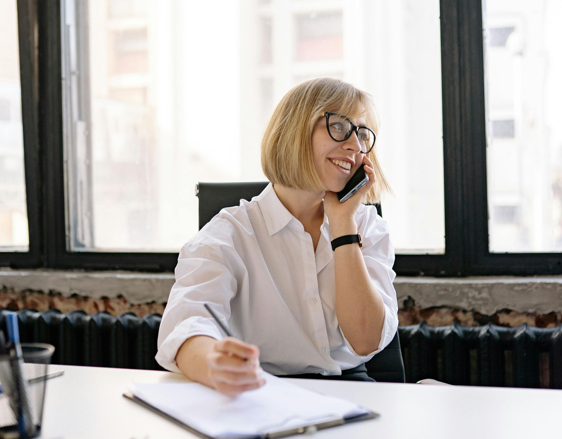 Person at a desk on the phone, smiling with a notebook in a bright office.