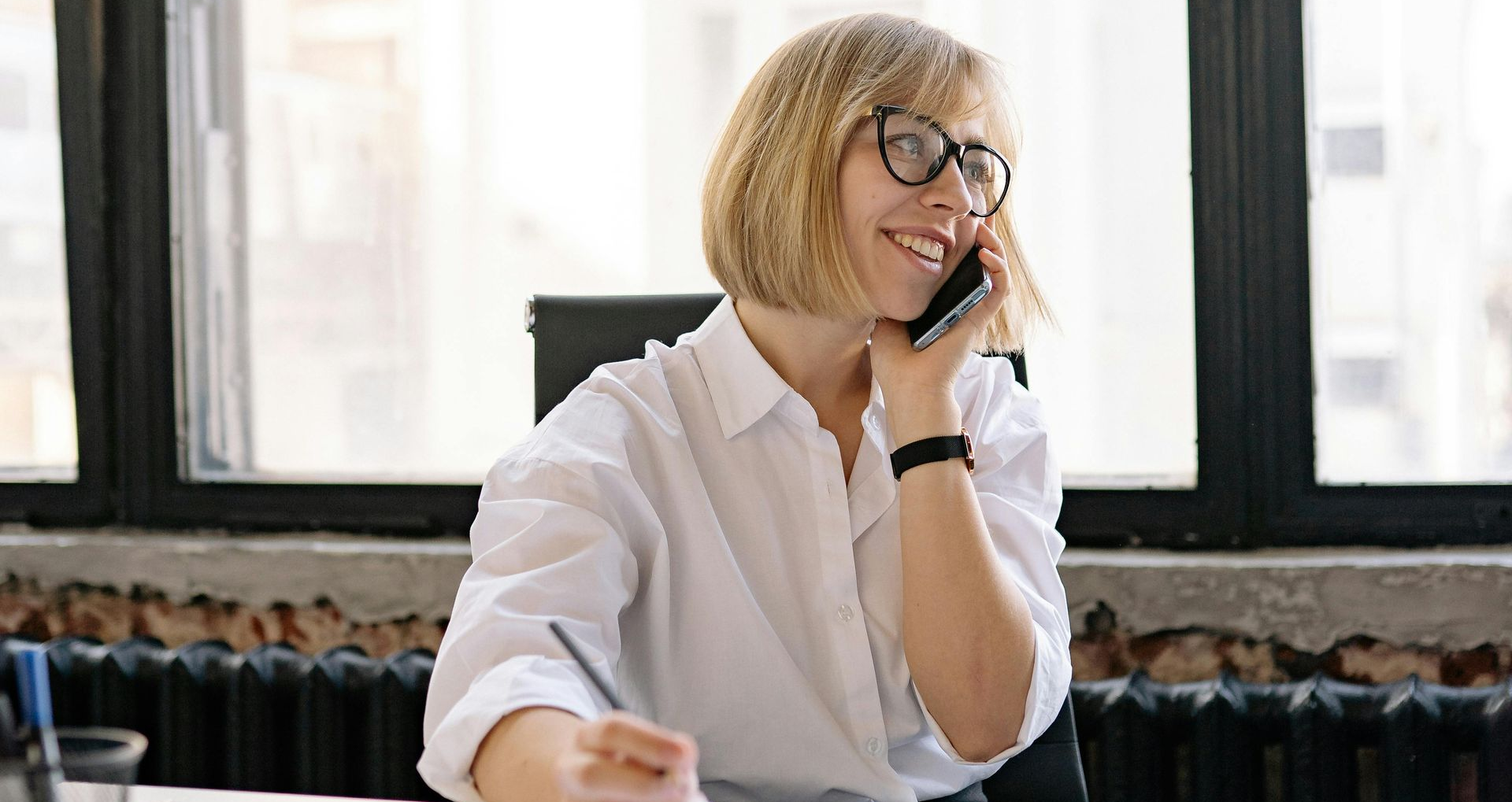 Person in glasses smiling at a desk in a bright office, holding a phone to their ear