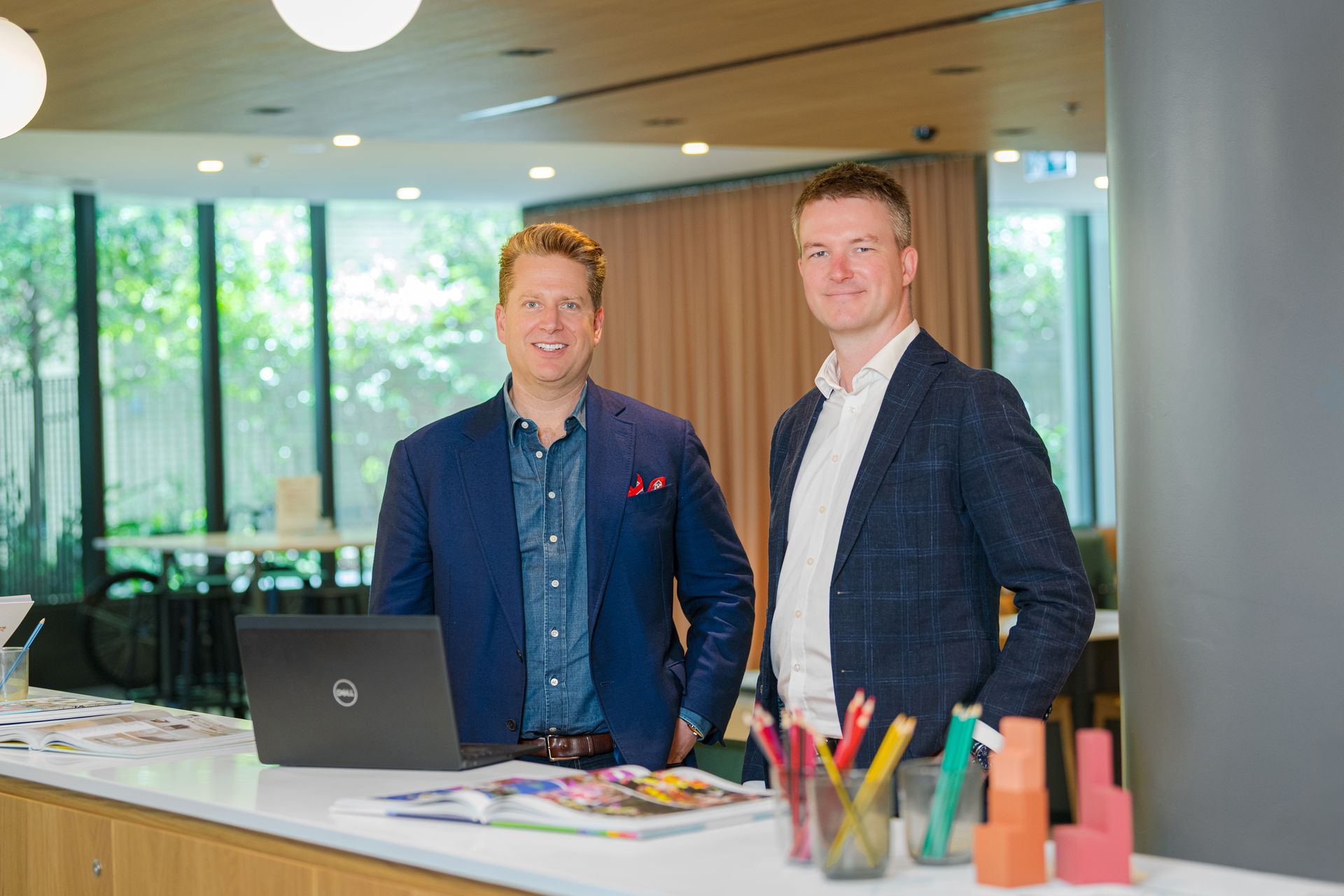 Two men stand at a white counter in a bright office lobby with a laptop and colorful cups nearby.