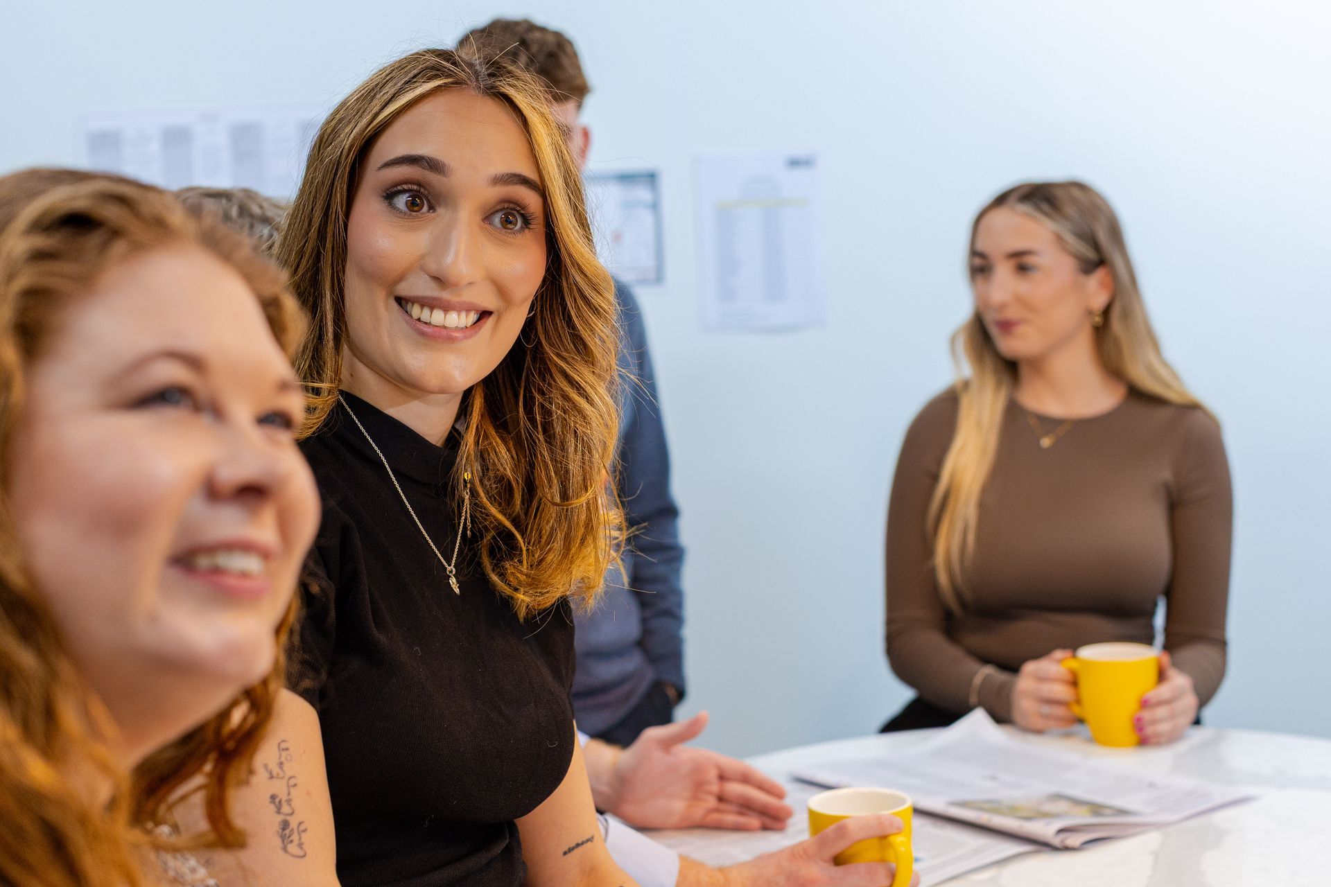 Three coworkers in a meeting, smiling around a table with papers and a yellow mug.