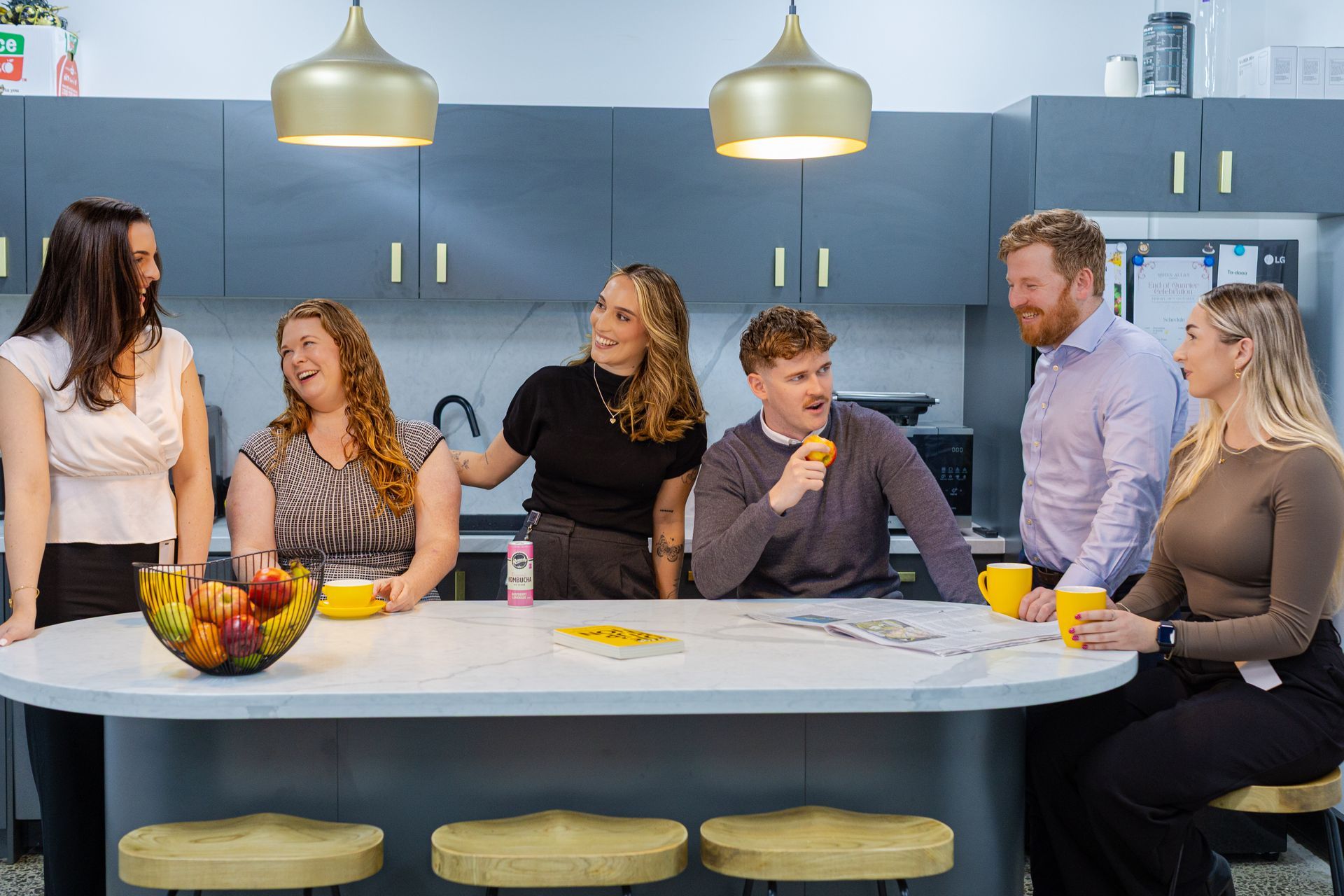 Six people chatting around a white kitchen island with fruit and drinks in a modern kitchen.
