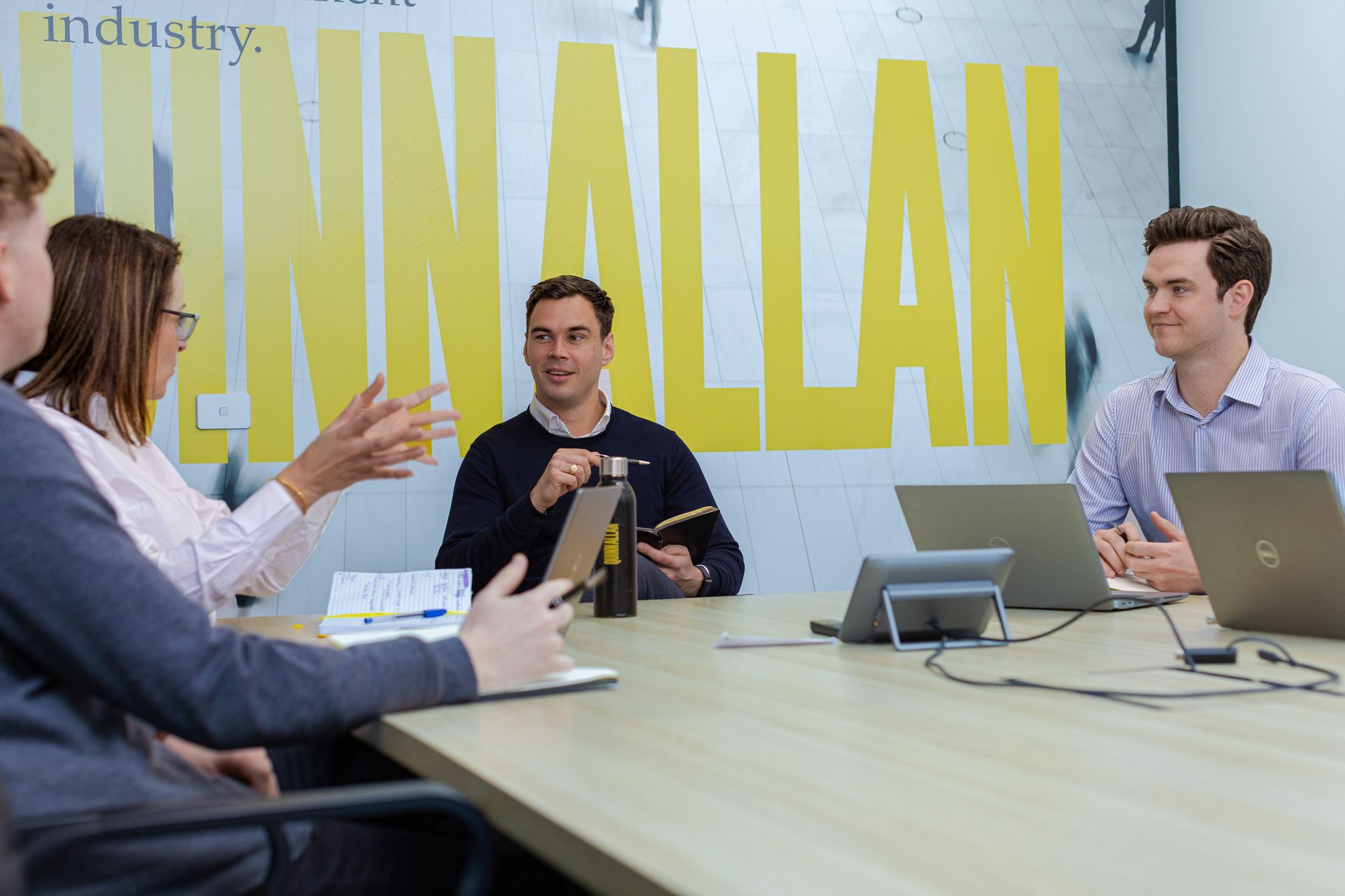 Business meeting in a bright office, with people around a table and a yellow wall reading “GERMANIAN.”
