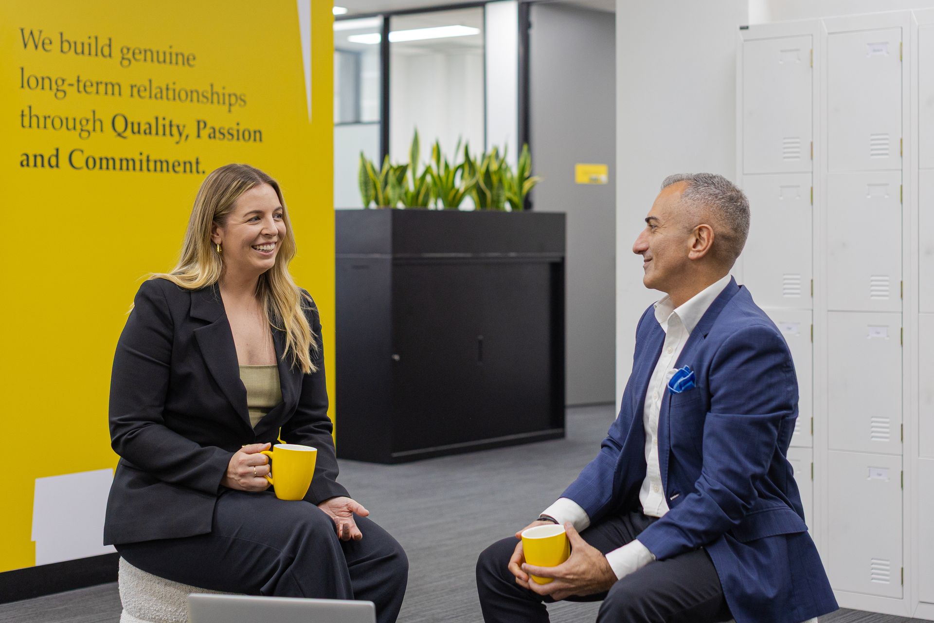 Two coworkers in suits chat over coffee in a modern office lounge with a yellow wall quote.