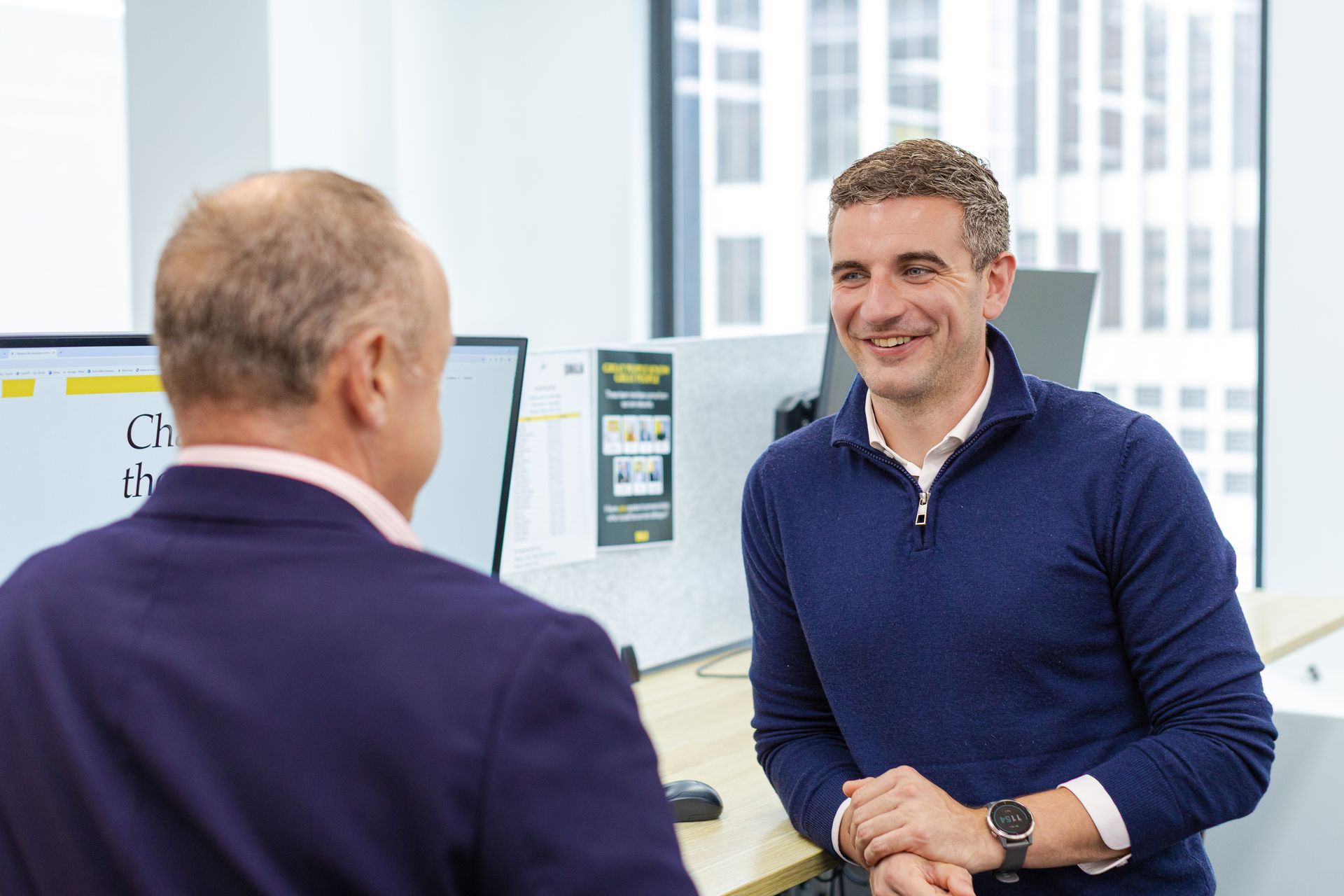 Two people chatting in a bright office, one smiling in a blue sweater and the other seen from behind in a purple blazer