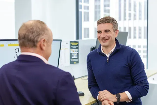 Two people talking in an office, one smiling and wearing a blue sweater.