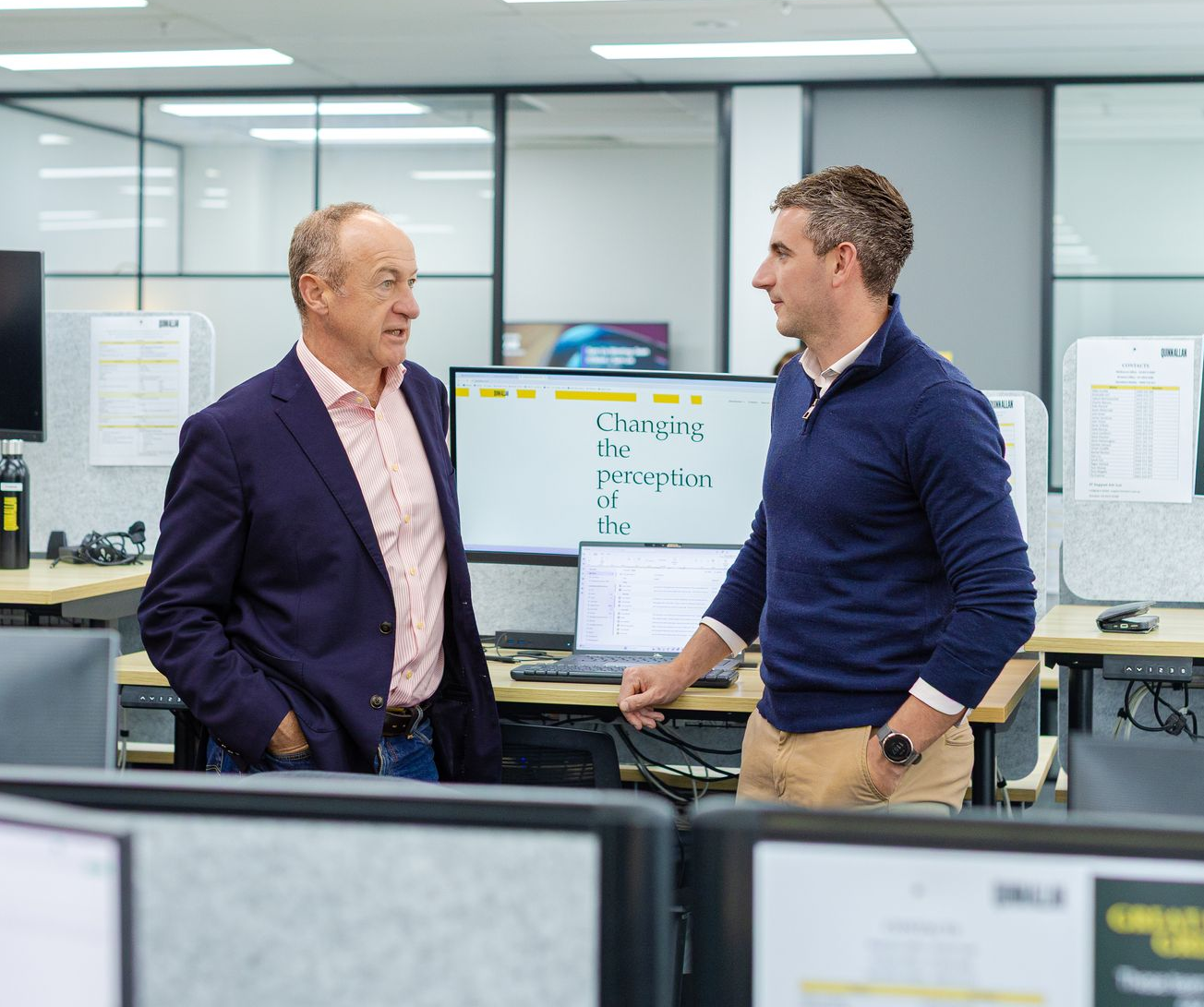 Two men talking in a modern office with computer monitors in the background.