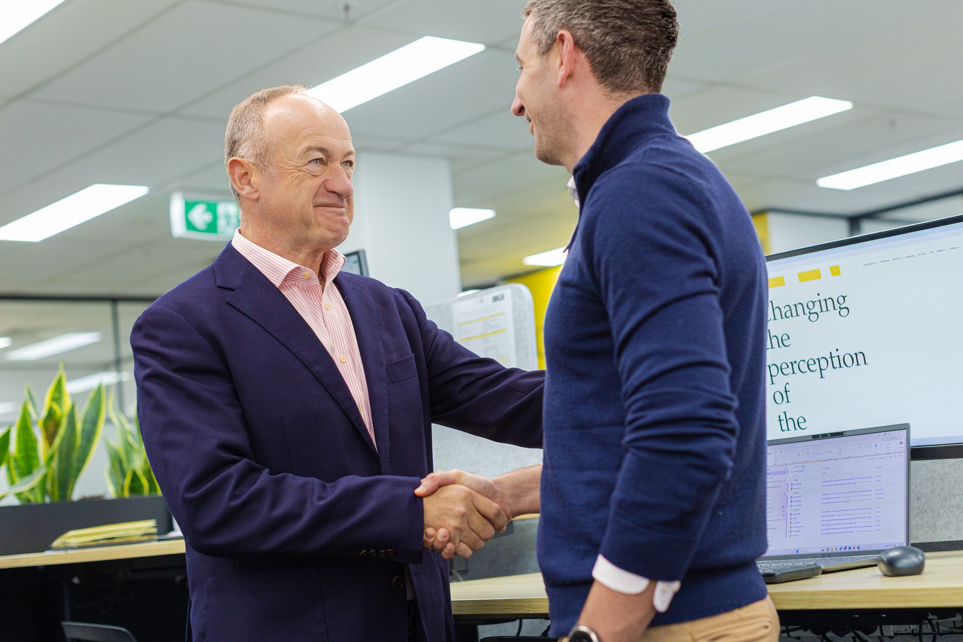 Two men in an office shaking hands, with a laptop and whiteboard in the background.