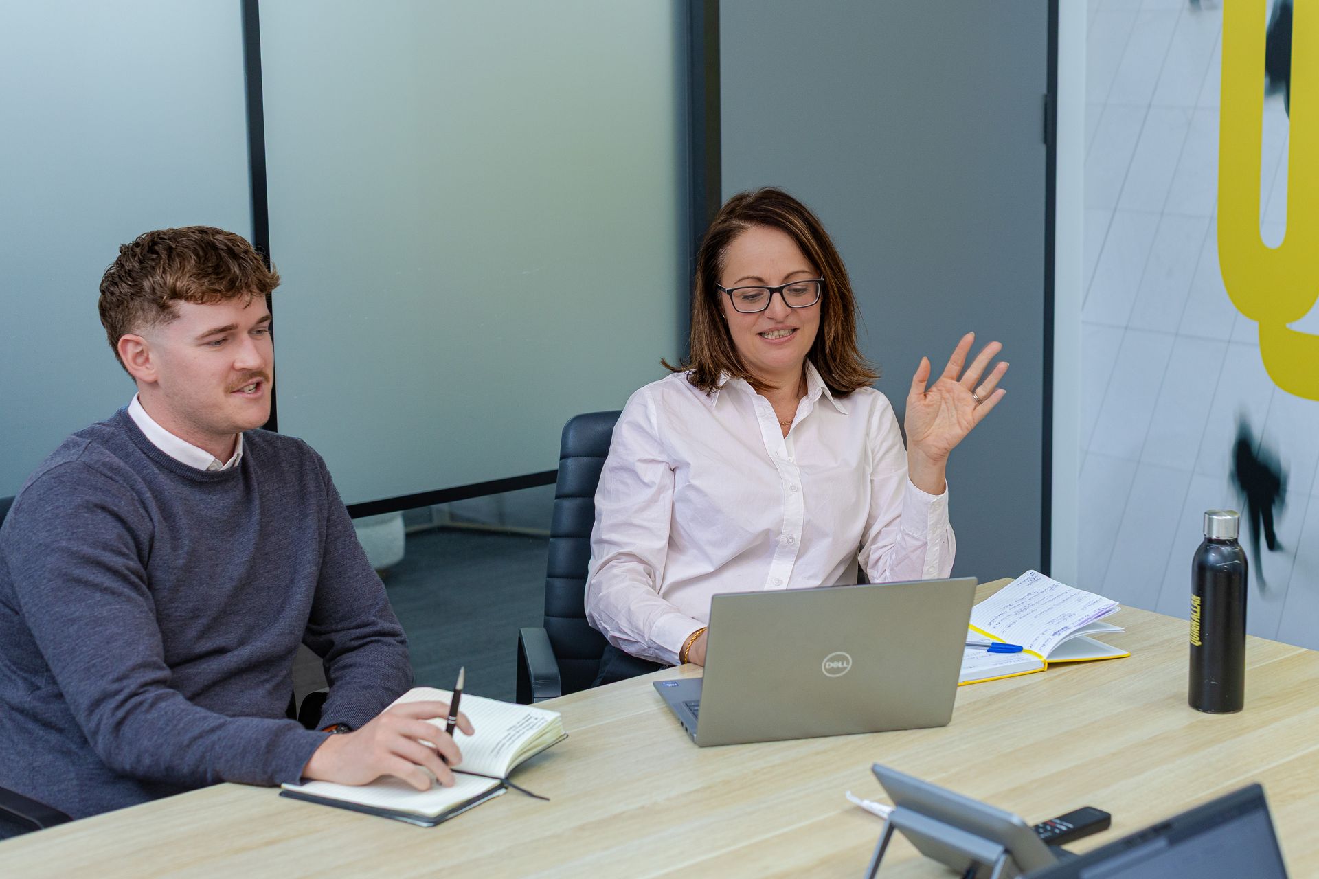 Two colleagues at a conference table; one gestures while the other takes notes beside a laptop.