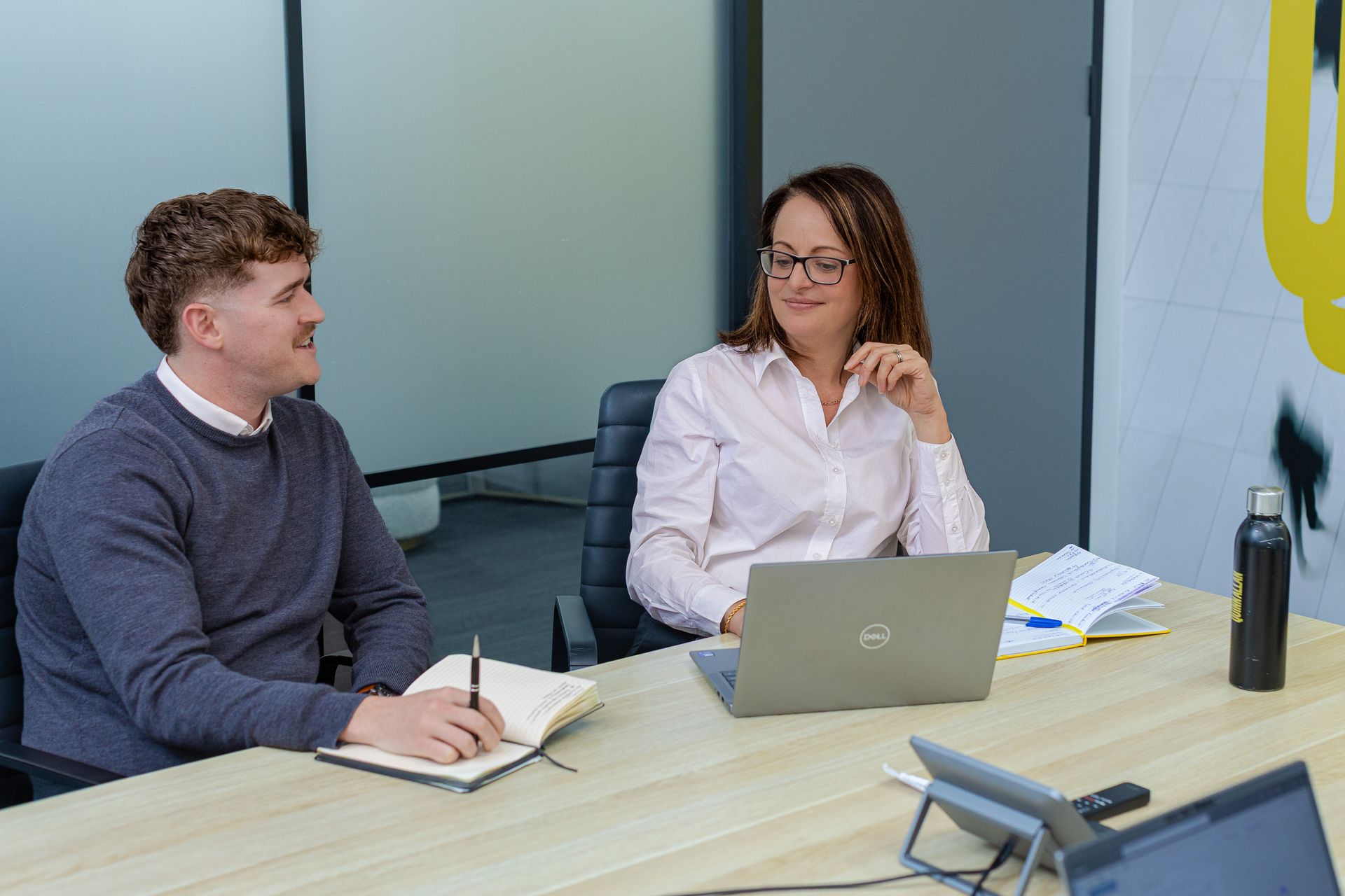 Two colleagues in a meeting at a conference table, one taking notes and one speaking beside a laptop.