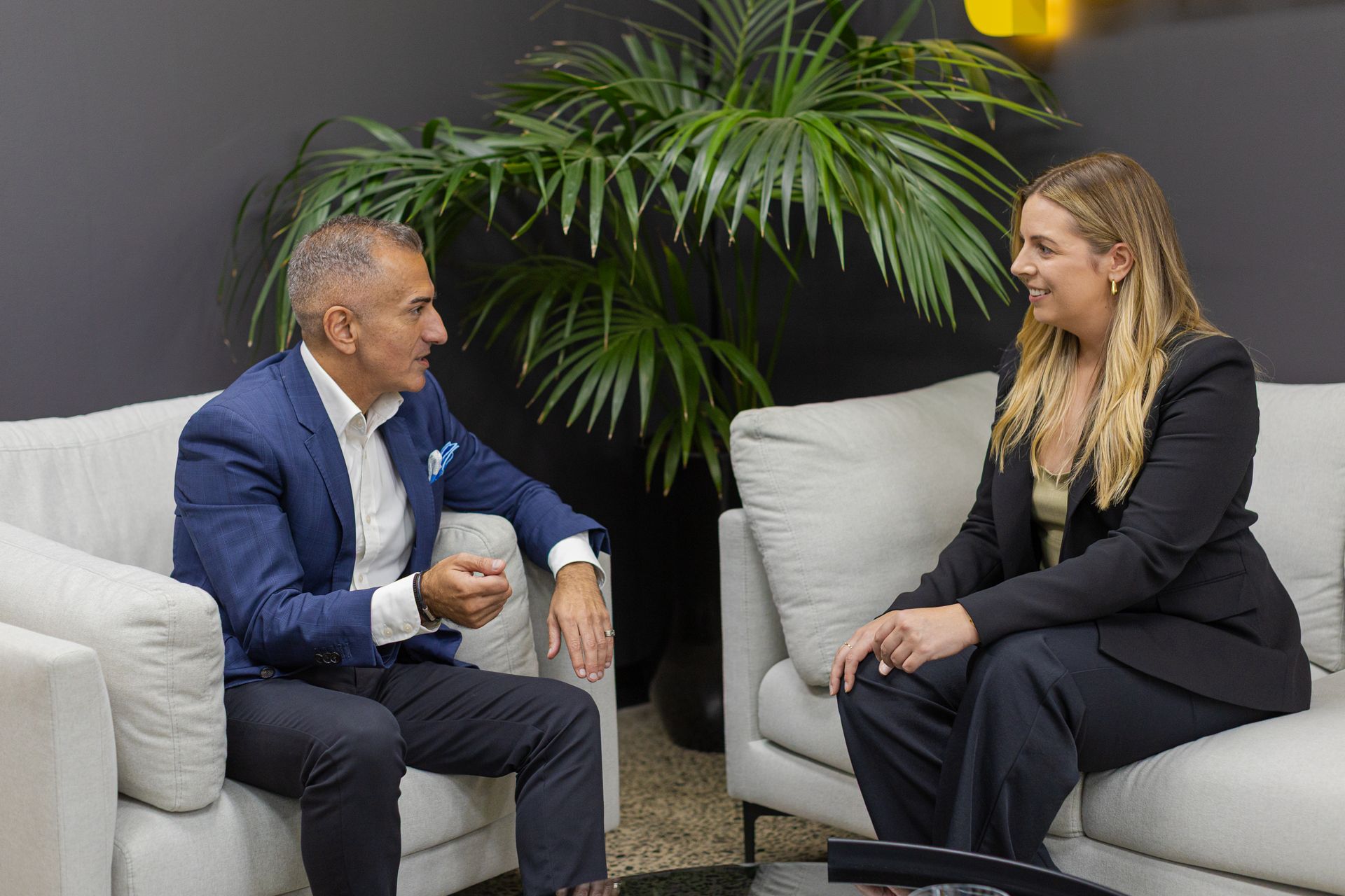 Two people in business attire talking on gray couches in an office lounge with a large plant behind them