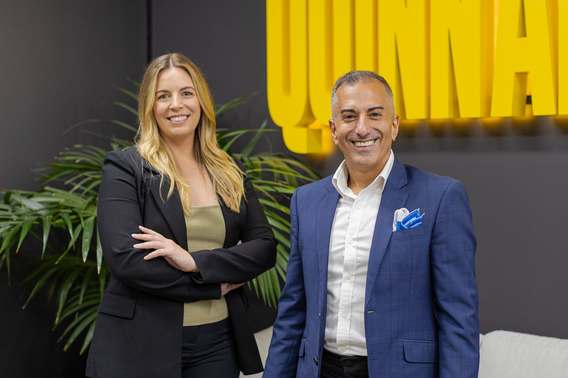 Two professionals posing in an office, with a yellow logo on the wall behind them.