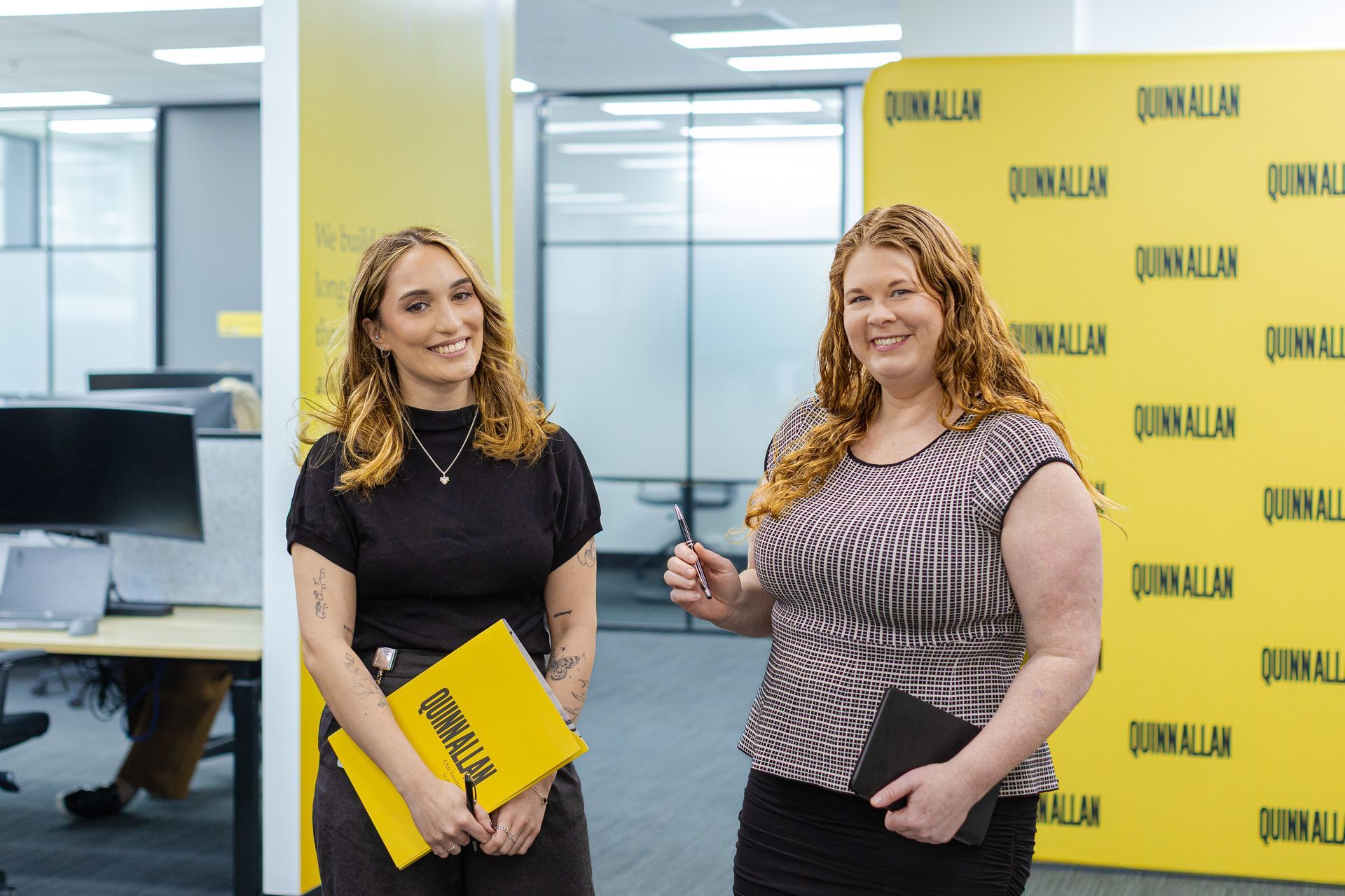 Two women stand in an office lobby, holding yellow folders in front of a yellow branded backdrop.